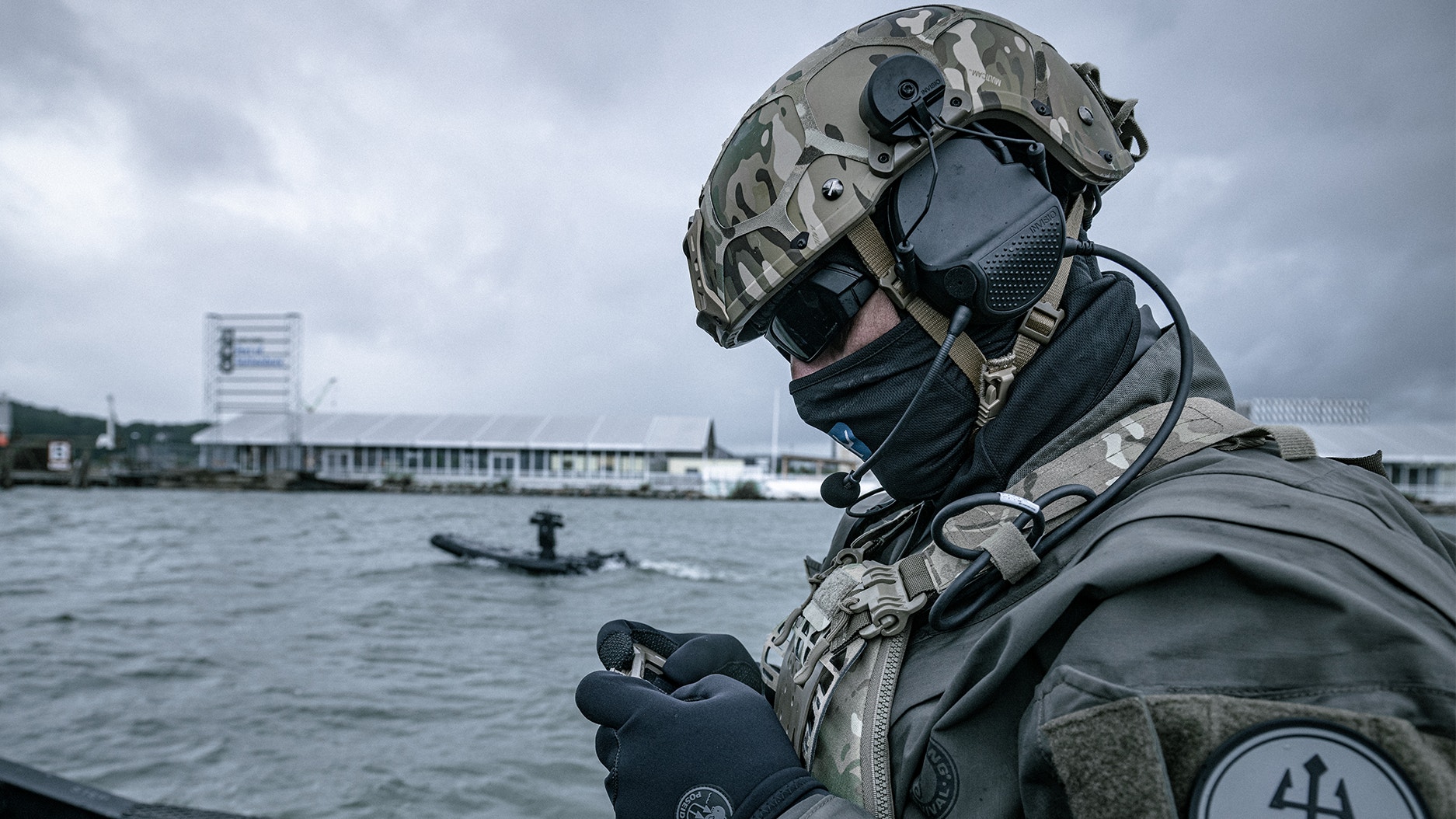 En kamouflageklädd soldat med hjälm och headset står på en båt vid en hamn, med en mindre båt synlig i vattnet bakom.