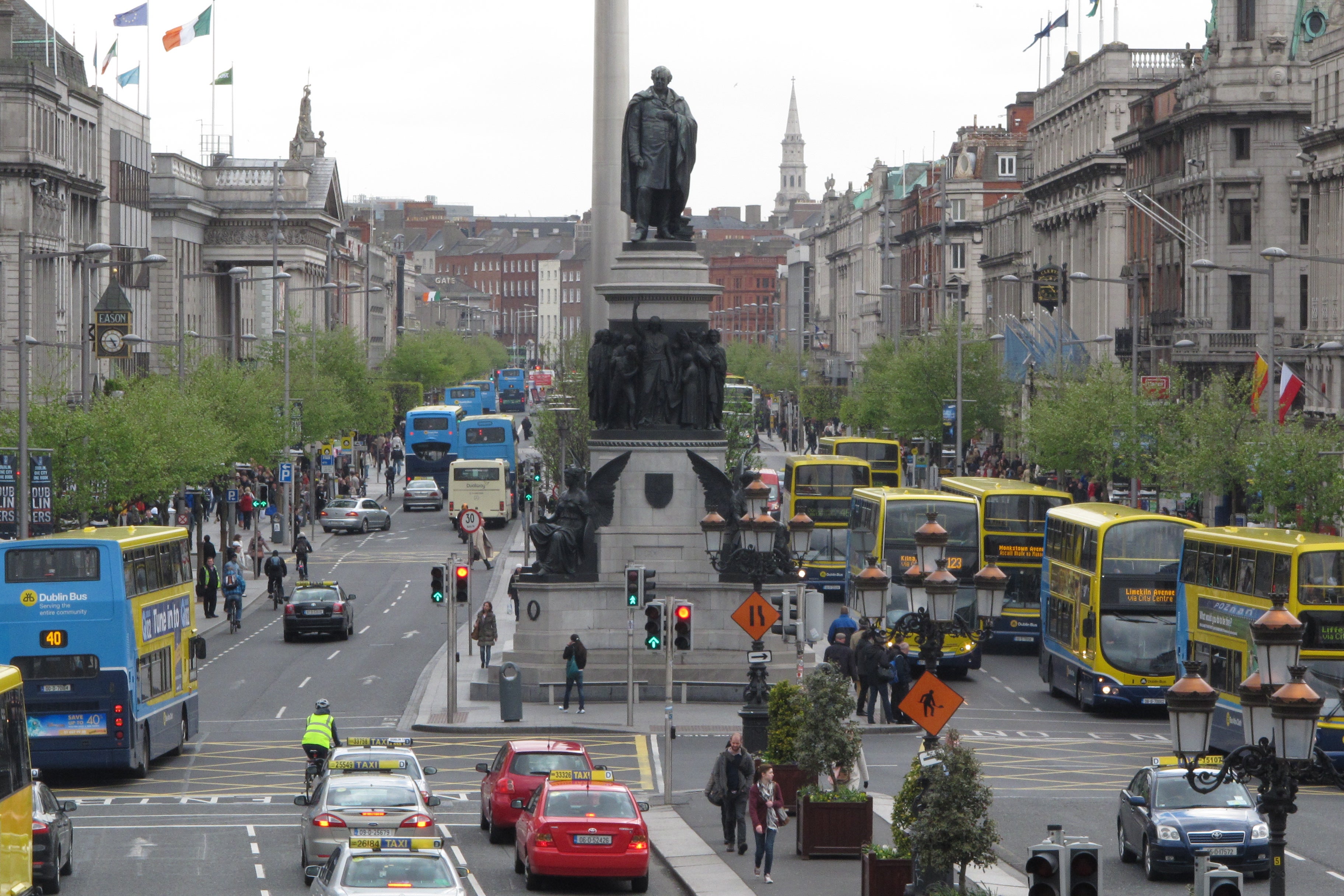 Livlig gata i Dublin med dubbeldäckarbussar, bilar och fotgängare runt en hög monumentstaty.