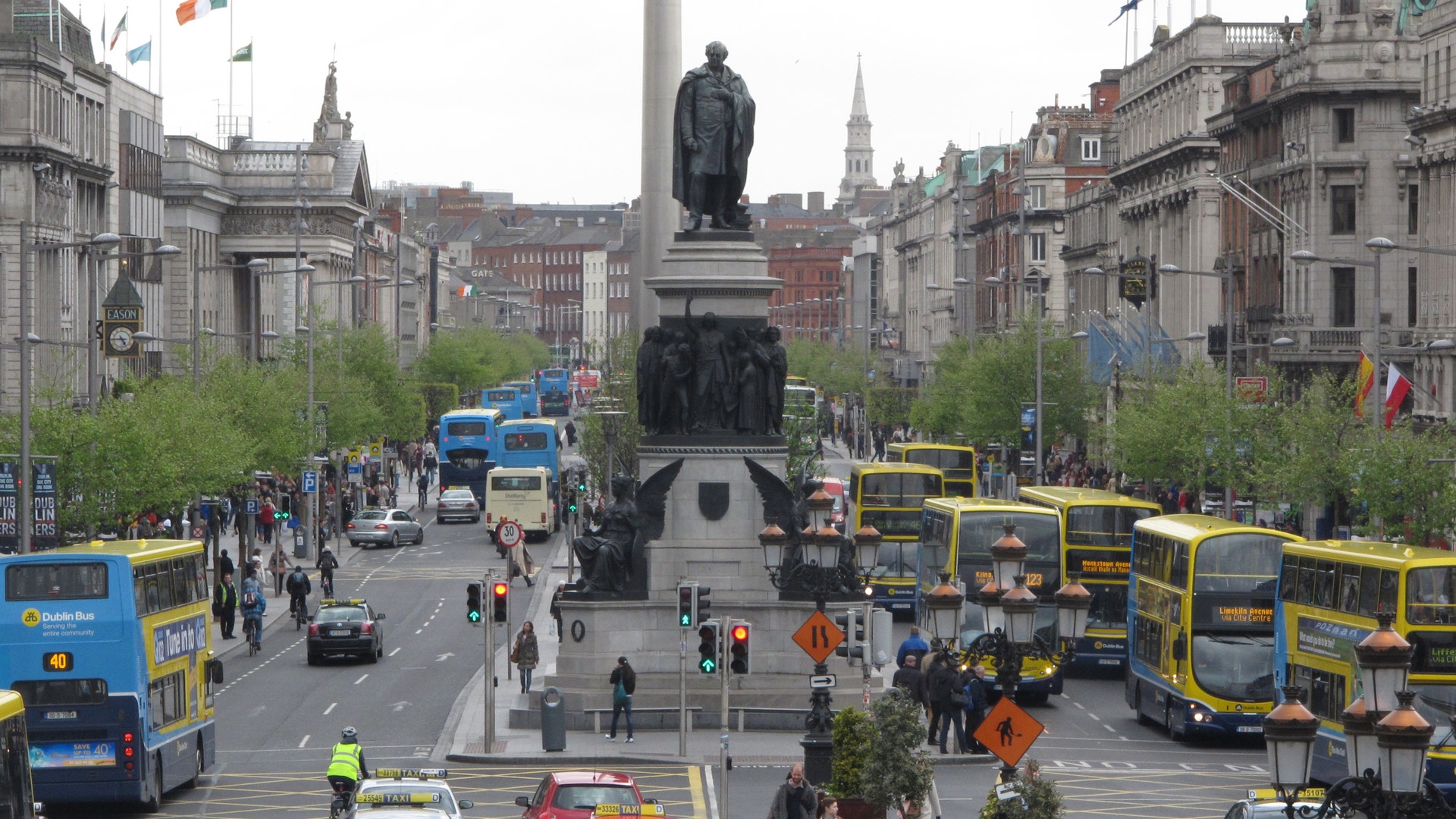 Livlig gata i Dublin med dubbeldäckarbussar, bilar och fotgängare runt en hög monumentstaty.