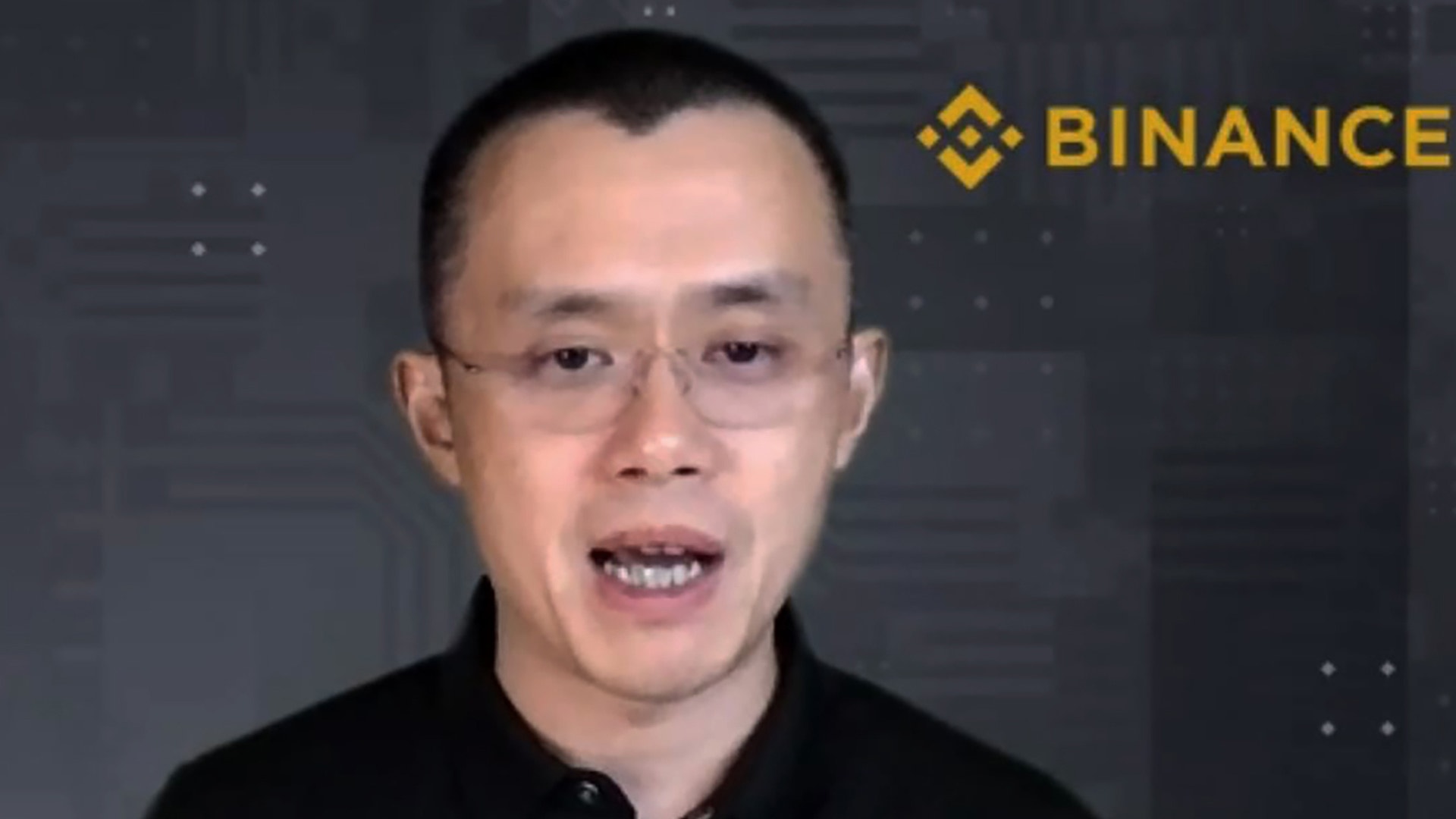 Porträtt av en man med kort mörkt hår, glasögon och svart tröja som talar in i kameran. I bakgrunden syns Binance-logotypen.