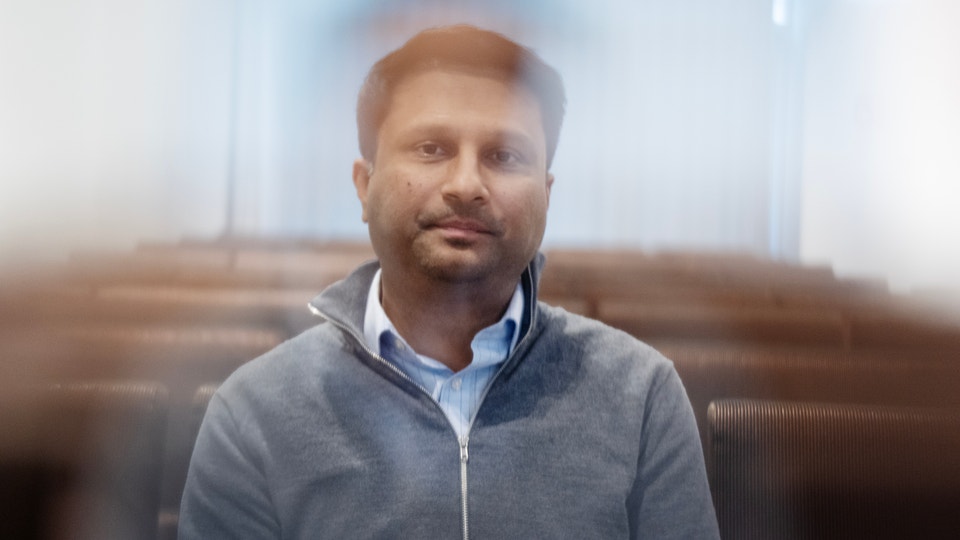 Porträtt av en man med kort brunt hår och skägg, klädd i en grå dragkedjetröja över en ljusblå skjorta. Han är fotograferad inomhus mot en suddig bakgrund med möbler, och tittar direkt mot kameran med ett lugnt uttryck.