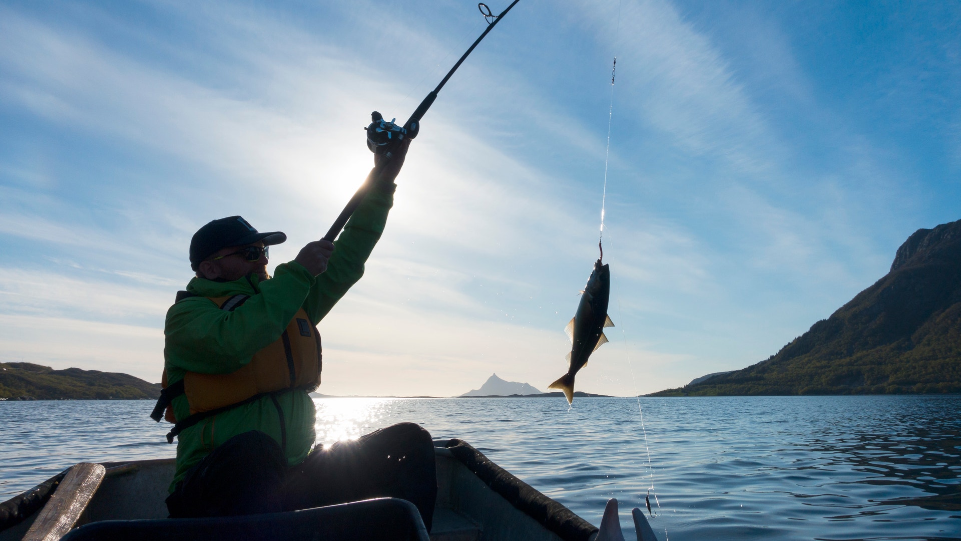 En person i en båt drar upp en fisk med ett fiskespö vid en fjord, i motljus under blå himmel.