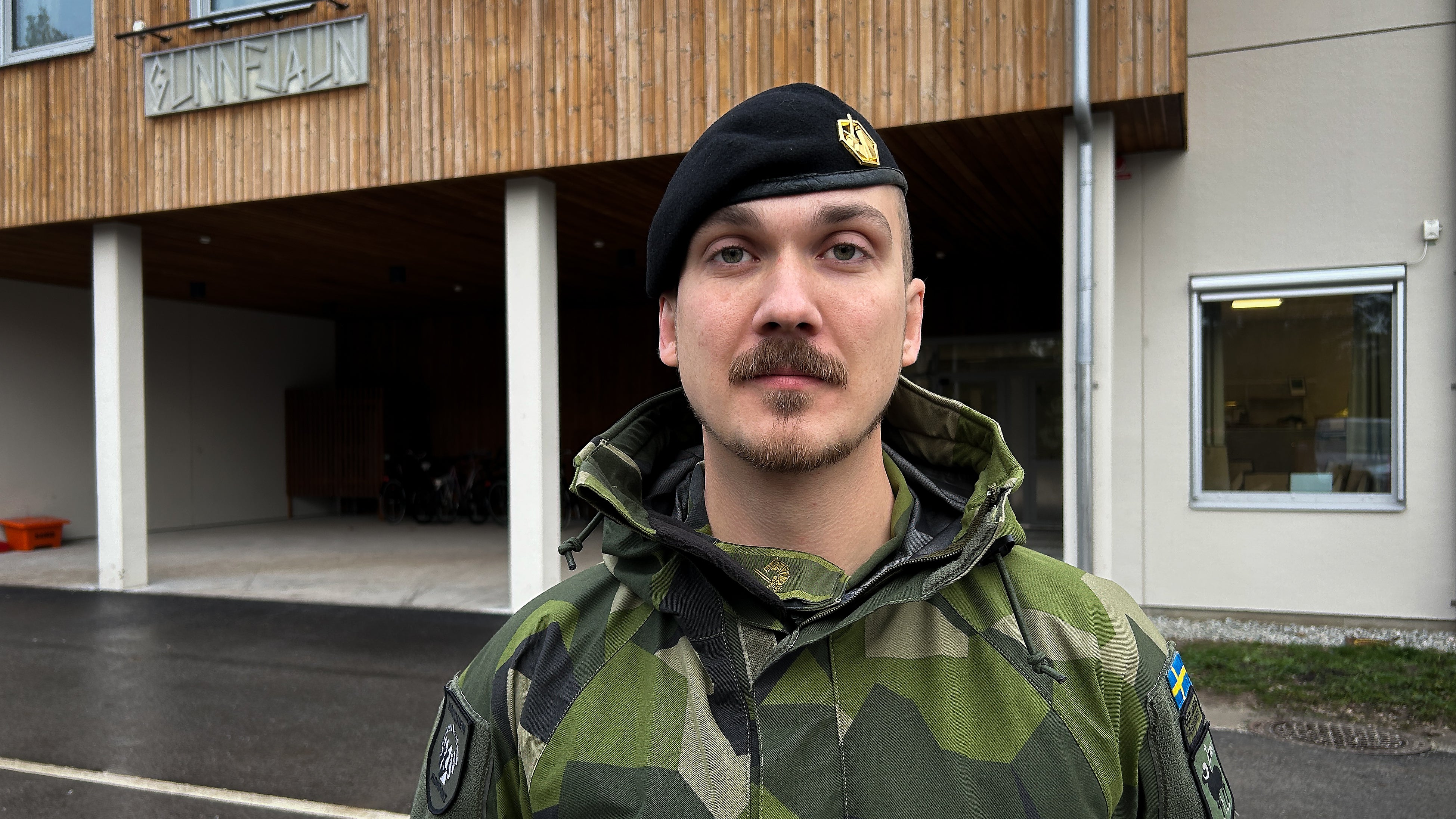En man i militär uniform med svart baskermössa och gröncamouflagejacka står framför en byggnad med träfasad.