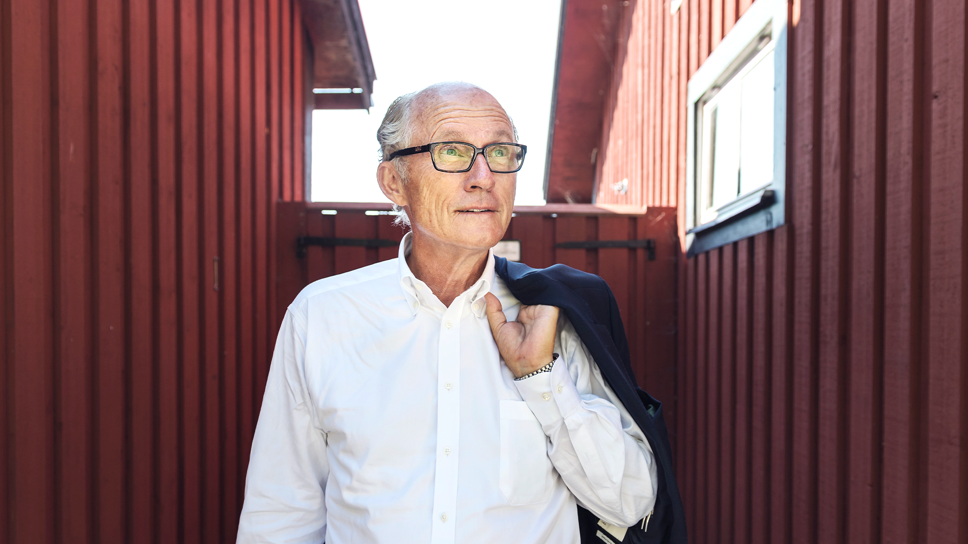 Äldre man med glasögon i vit skjorta framför röd metallbyggnad