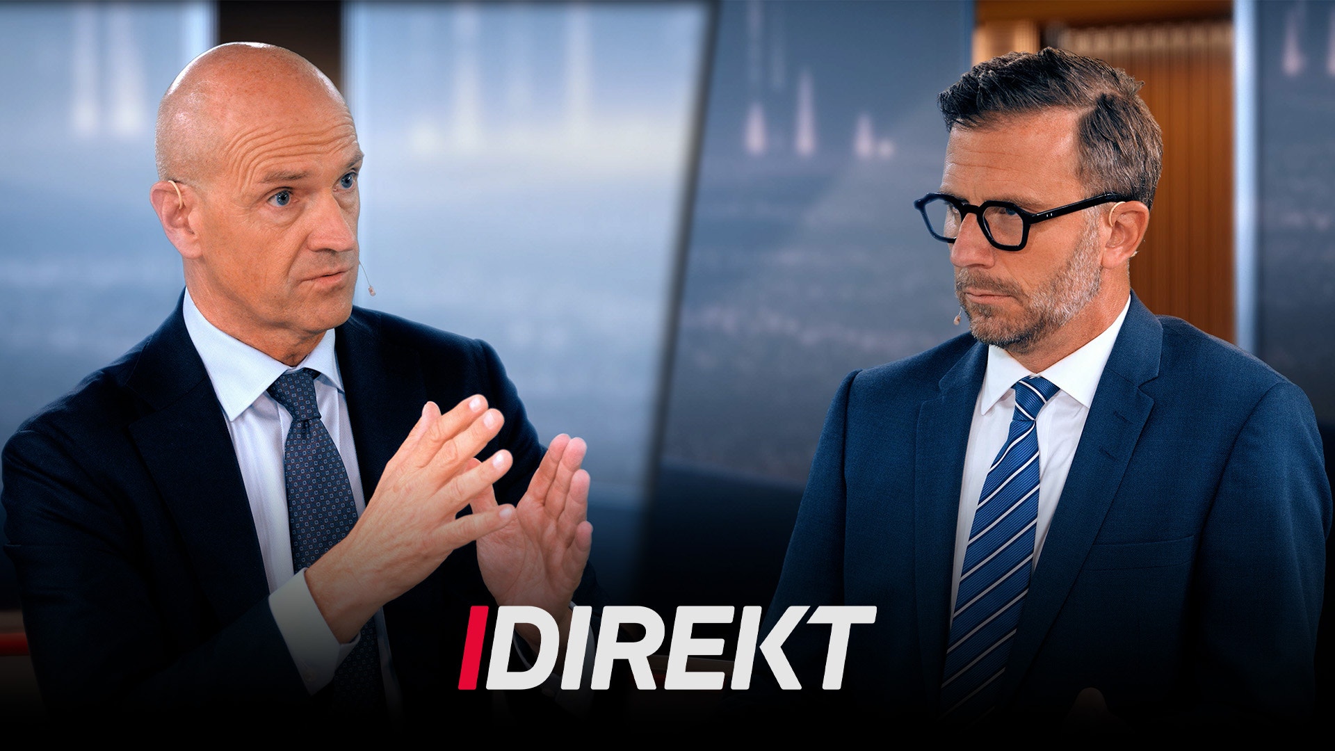 Två män i affärskläder sitter och diskuterar i ett modernt kontor med stadsvy i bakgrunden.