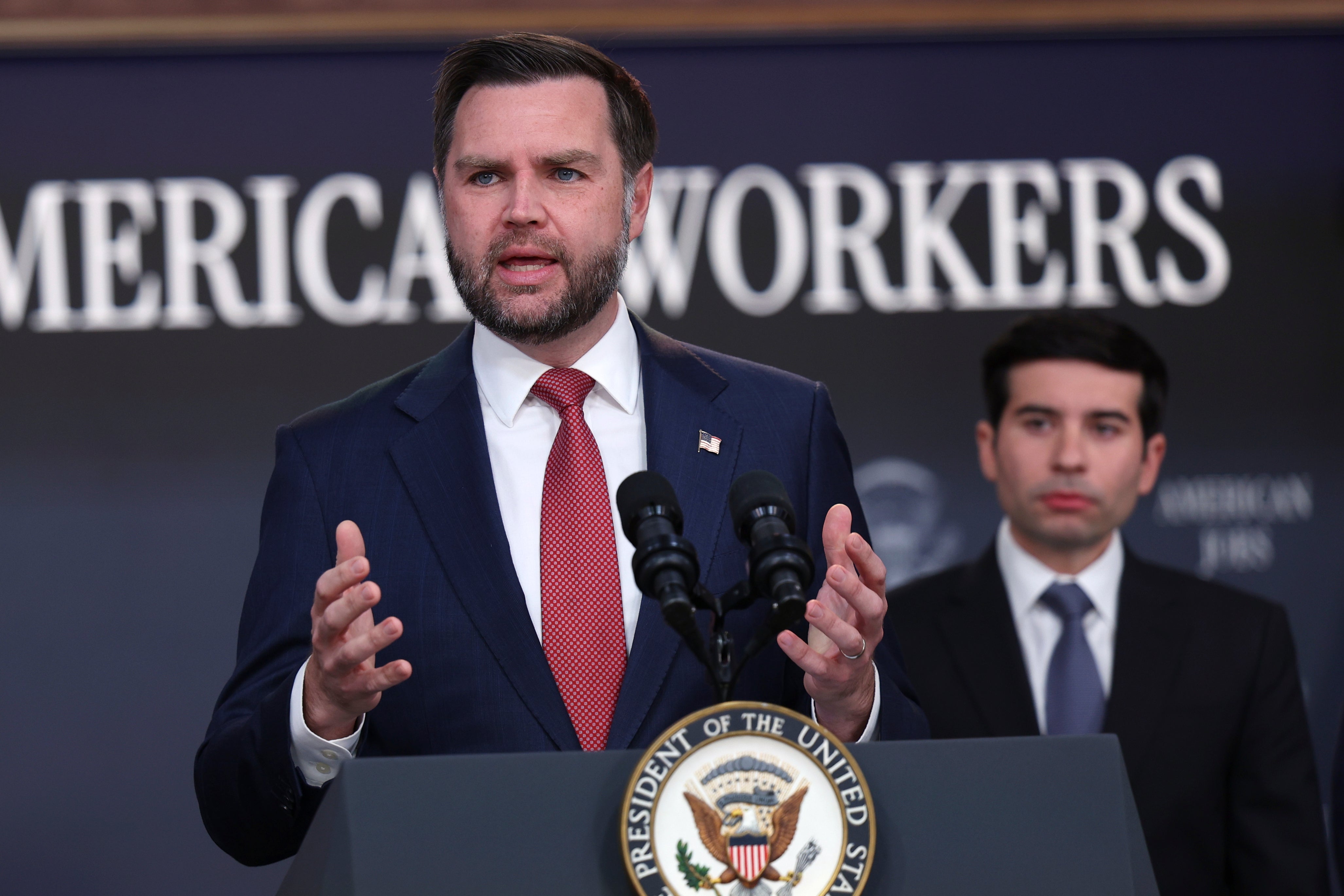 En man i kostym och slips talar vid en talarstol med USA:s vicepresidentsigill. Bakom honom står en annan man i kostym och lyssnar