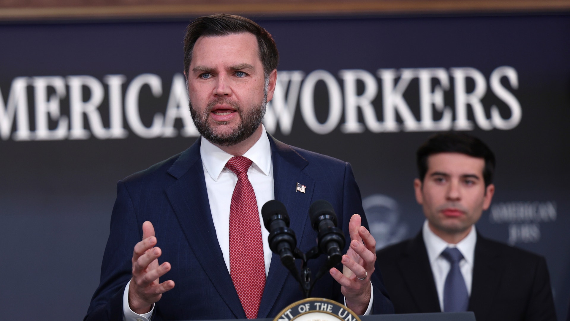 En man i kostym och slips talar vid en talarstol med USA:s vicepresidentsigill. Bakom honom står en annan man i kostym och lyssnar