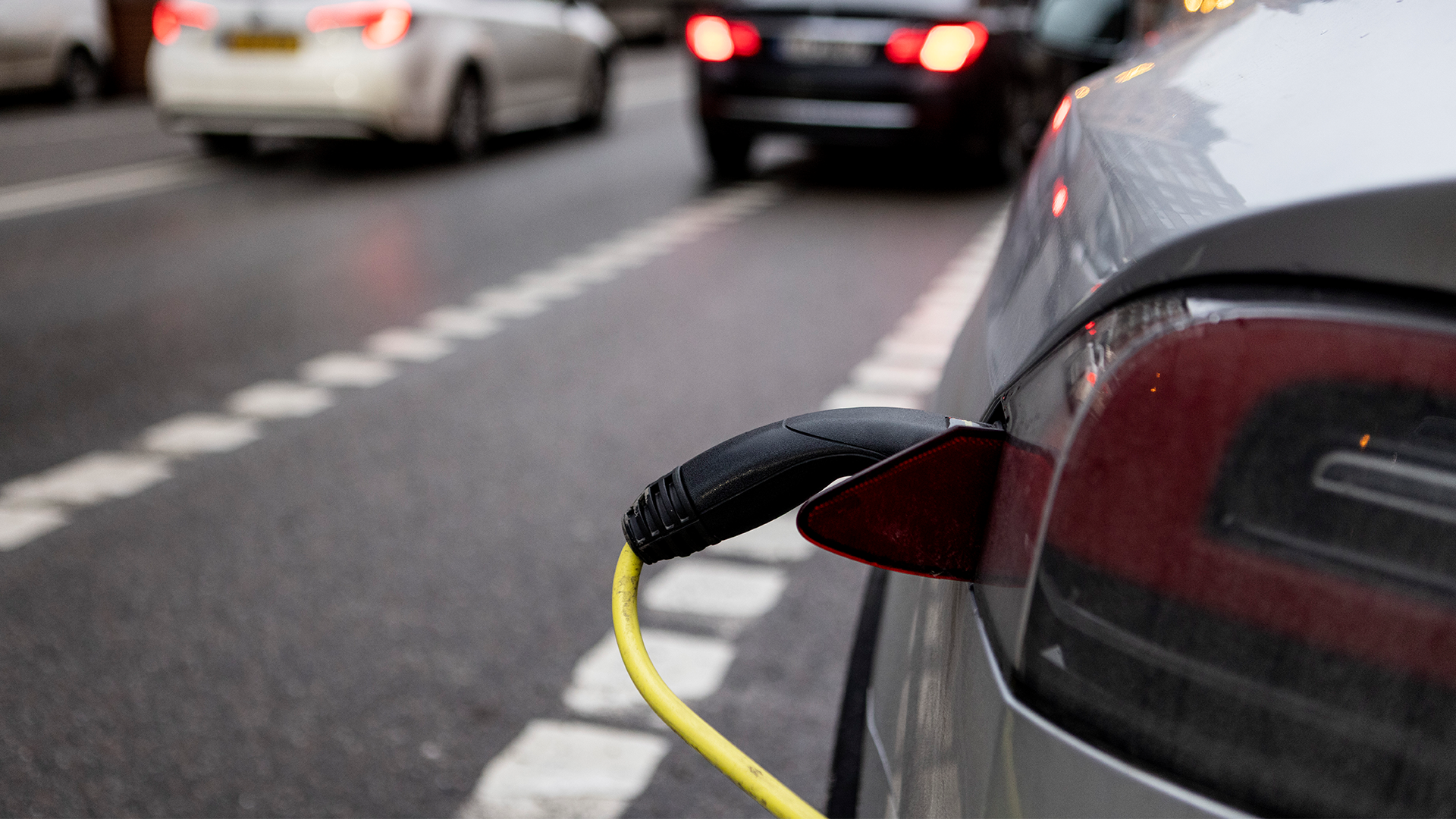 En röd elektrisk bil som laddas med en gul och svart laddkabel på en väg med andra fordon i bakgrunden.