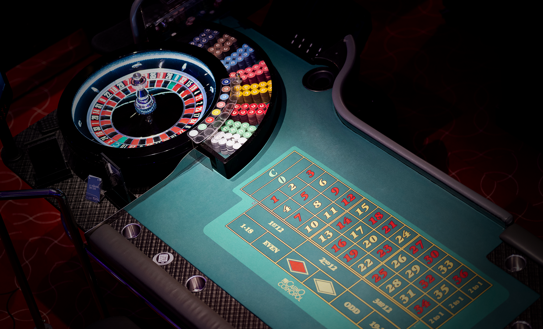 Livecasino-bord med ett klassiskt rouletthjul med röda och svarta nummer, samt ett grönt spelplan framför det. Hjulet är belyst med färgglada LED-lampor i blått, rött och gult. Spelplanen visar olika spelalternativ med nummer och symboler för betting.
