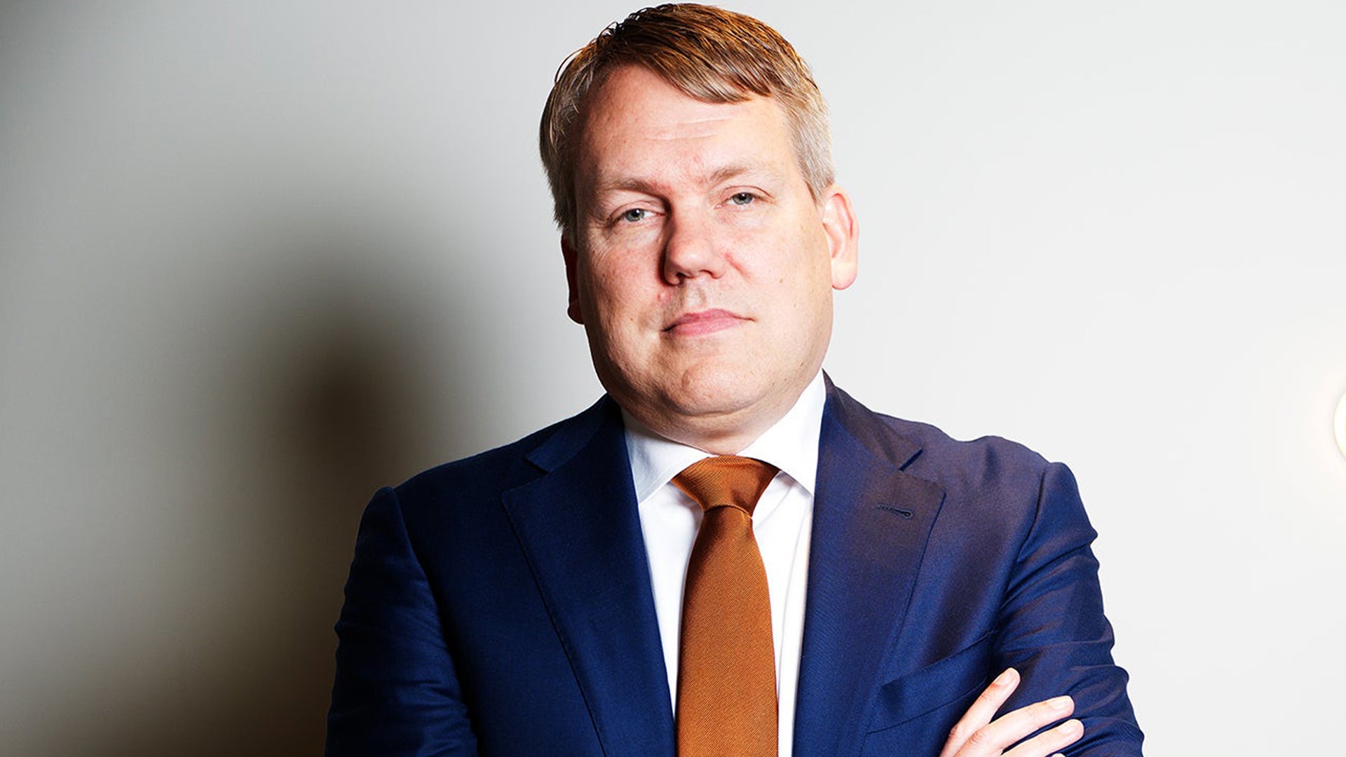 Porträtt av man i mörkblå kostym och orange slips mot ljus bakgrund.