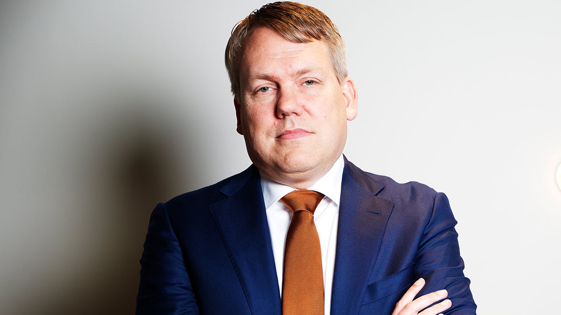 Porträtt av man i mörkblå kostym och orange slips mot ljus bakgrund.