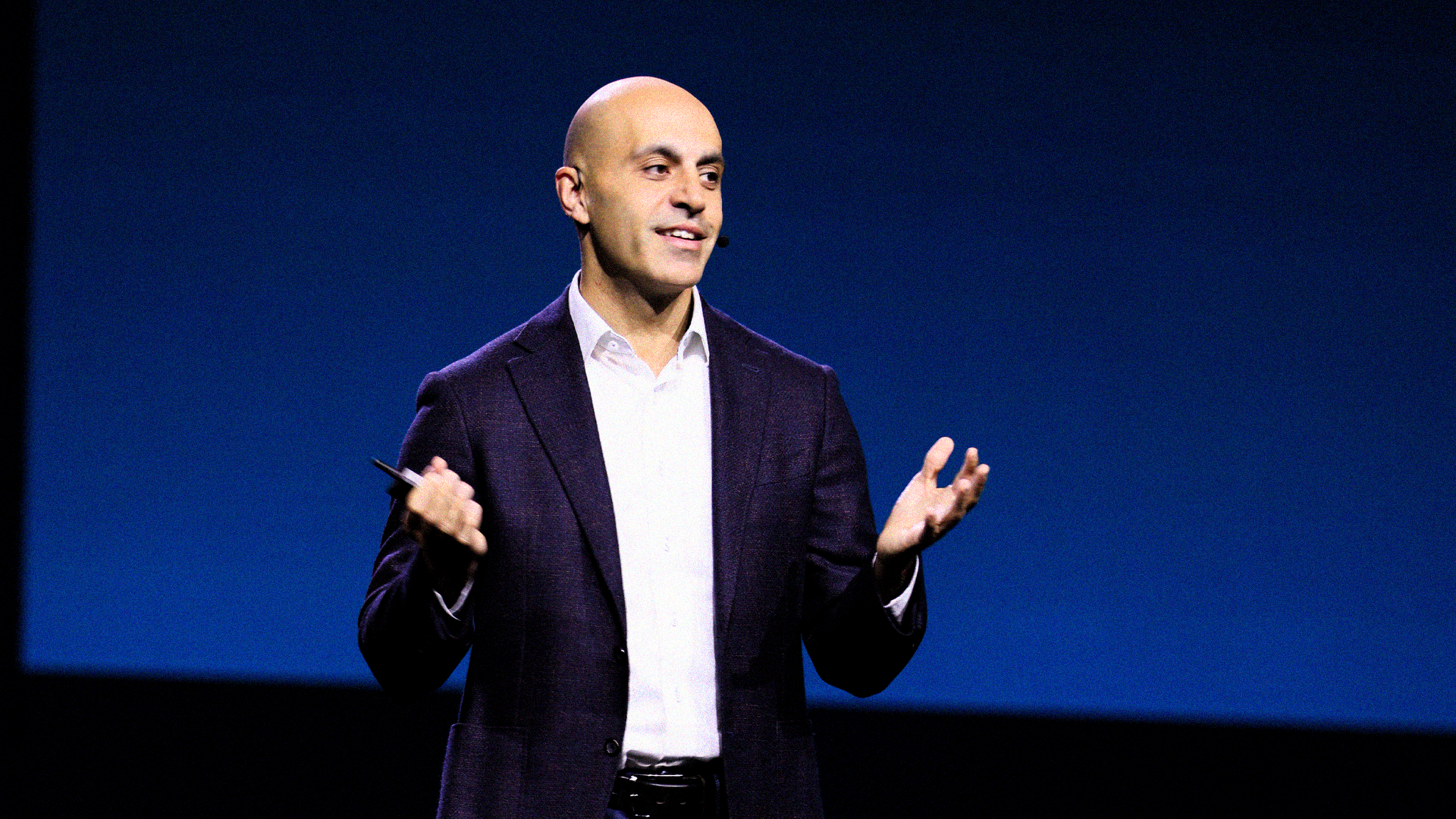 En man i mörkblå kavaj och vit skjorta presenterar på en scen med blå bakgrund.