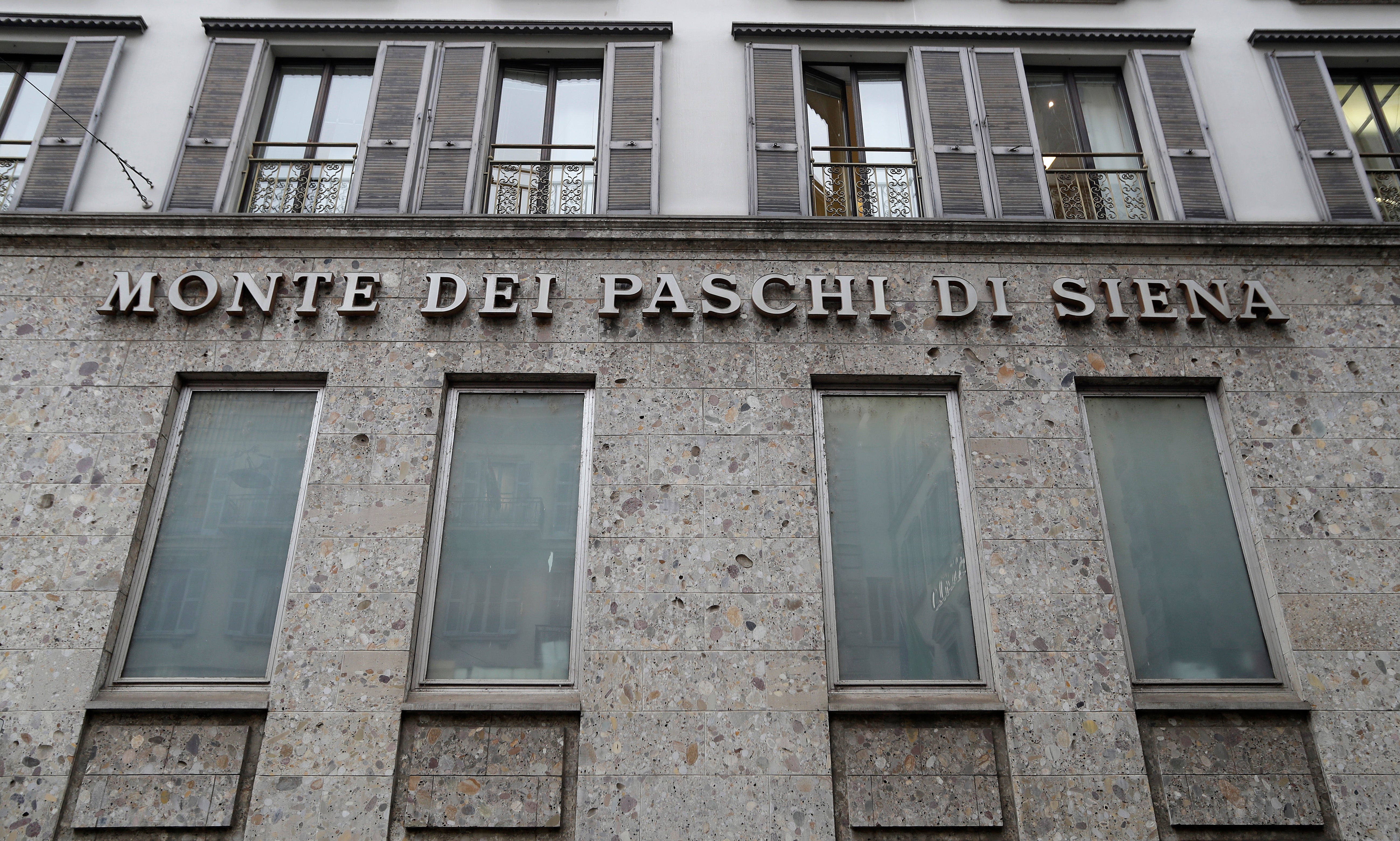 Fasad på en klassisk byggnad i sten med texten 'Monte dei Paschi di Siena' graverad på väggen. Byggnaden har flera höga rektangulära fönster och järnbalkonger på översta våningen.