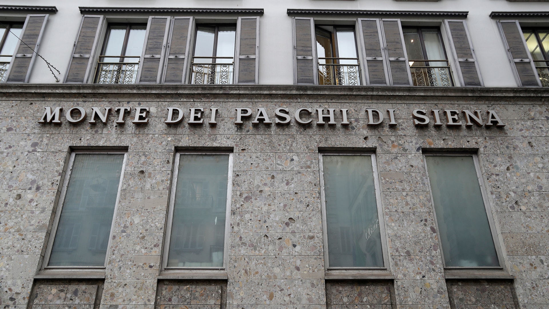 Fasad på en klassisk byggnad i sten med texten 'Monte dei Paschi di Siena' graverad på väggen. Byggnaden har flera höga rektangulära fönster och järnbalkonger på översta våningen.