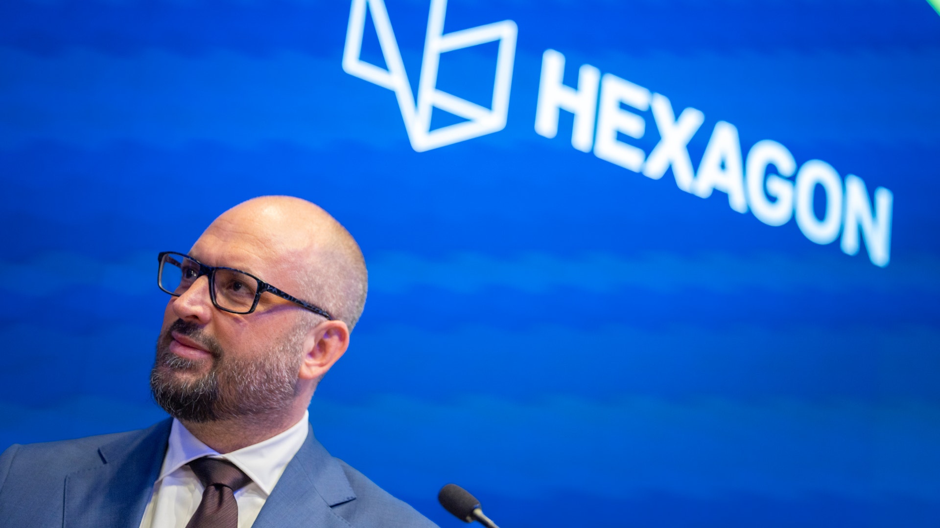 Man med glasögon och skägg talar vid en mikrofon framför en blå bakgrund med Hexagon-logotypen.,