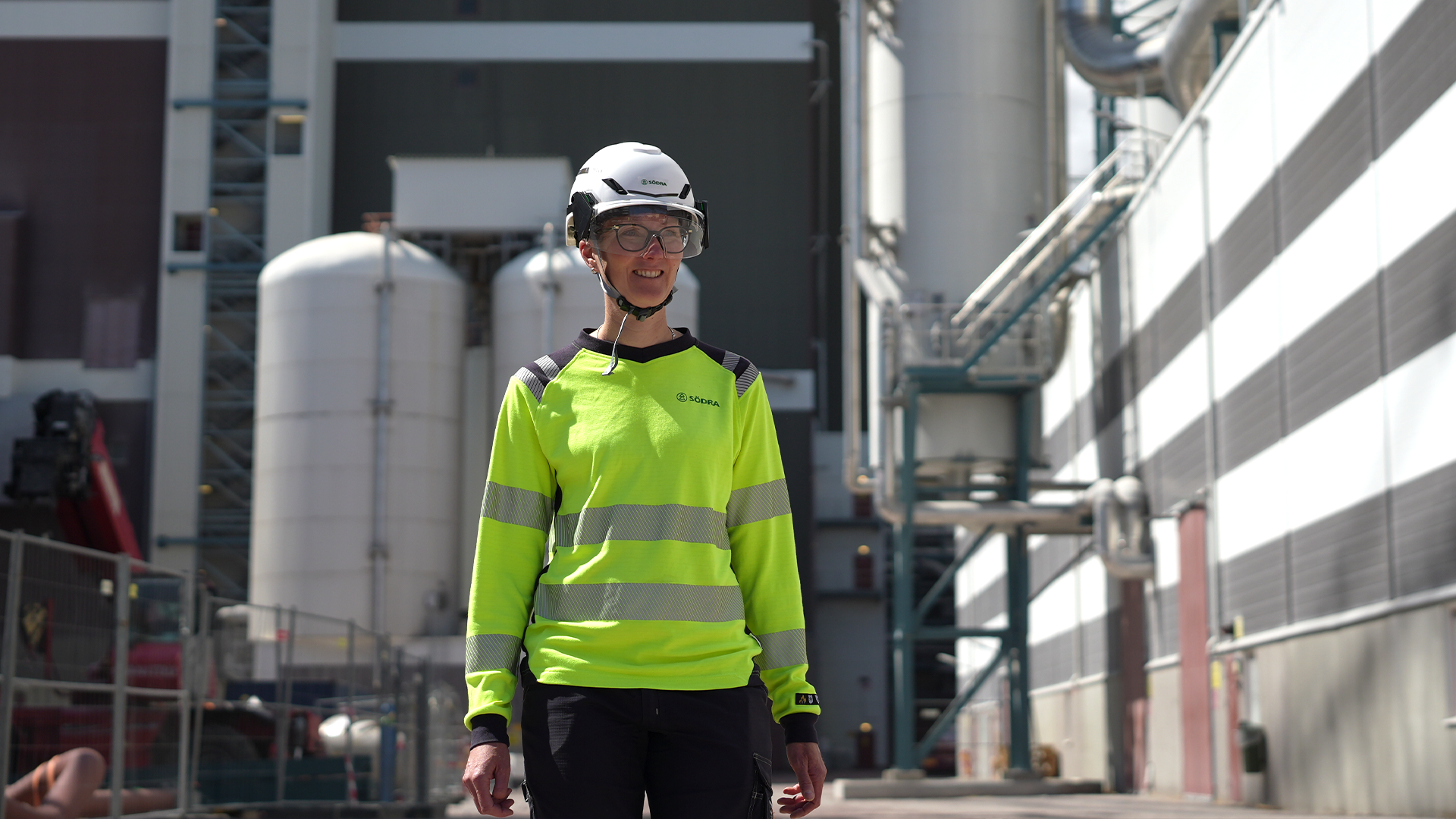 Arbetare i gul reflekterande väst och vit säkerhetshjalm på en industriell arbetsplats med metallstrukturer och tankar i bakgrunden.