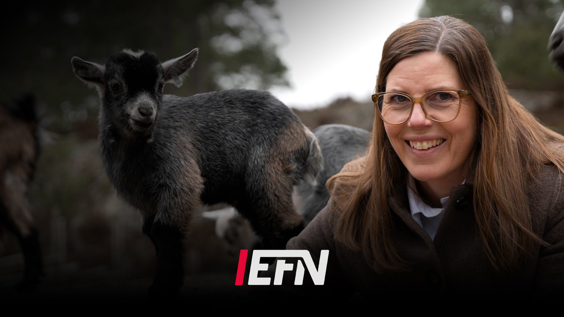 En kvinna ler mot kameran med en get bakom henne i en utomhusmiljö.