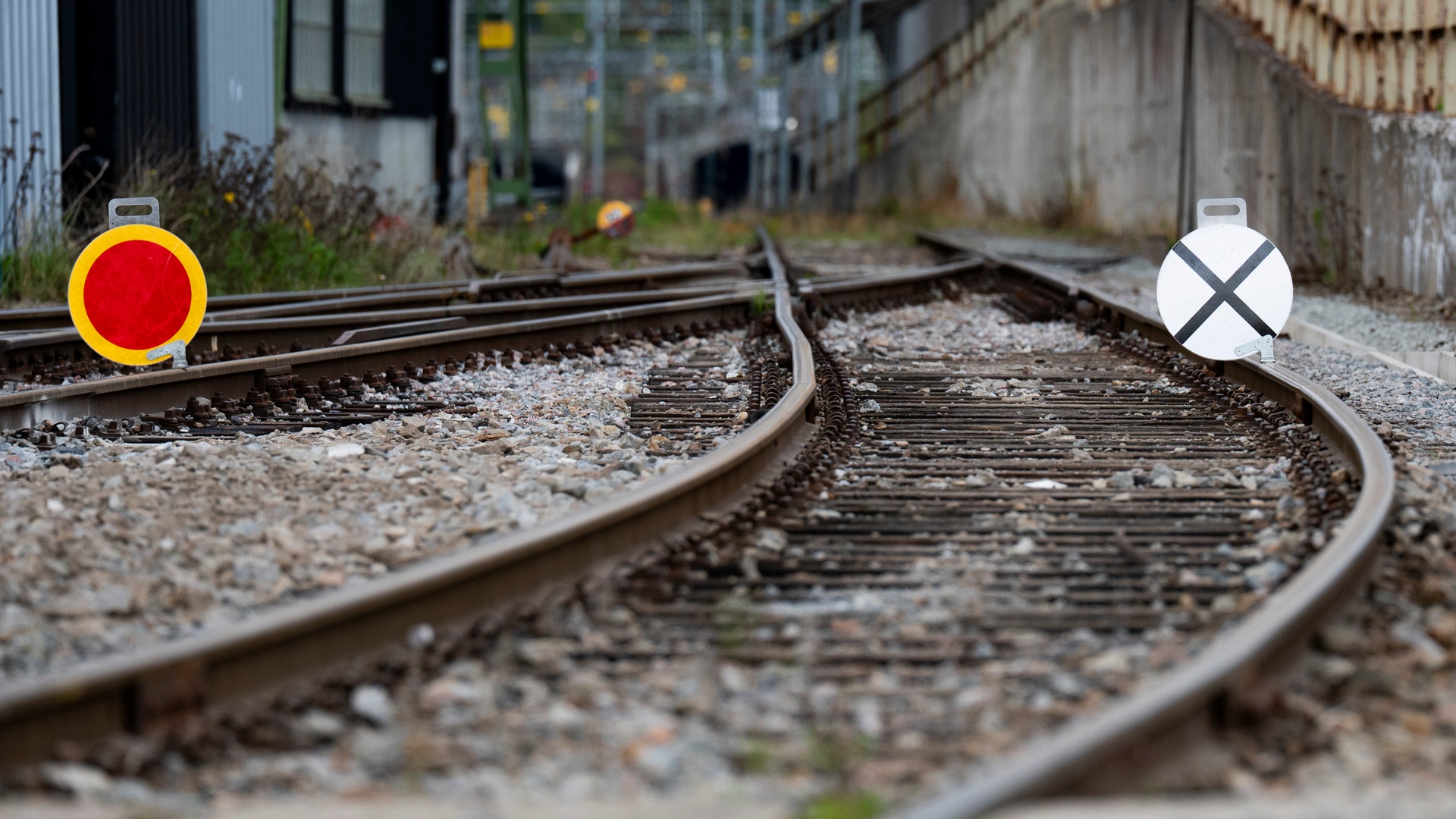Två järnvägsspår som korsar varandra. Det vänstra spåret är markerat med en röd och gul cirkel, det högra med ett vitt kryss. Urbana byggnader syns i bakgrunden.