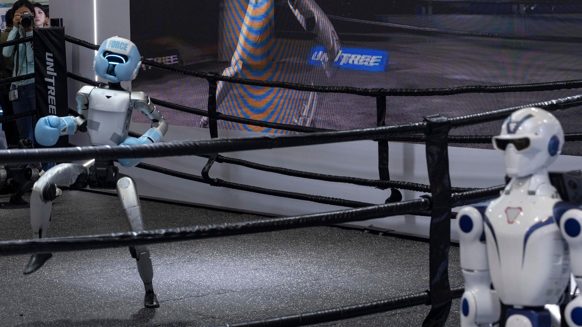 Två humanoidrobotar boxas mot varandra i en boxningsring med svarta rep.