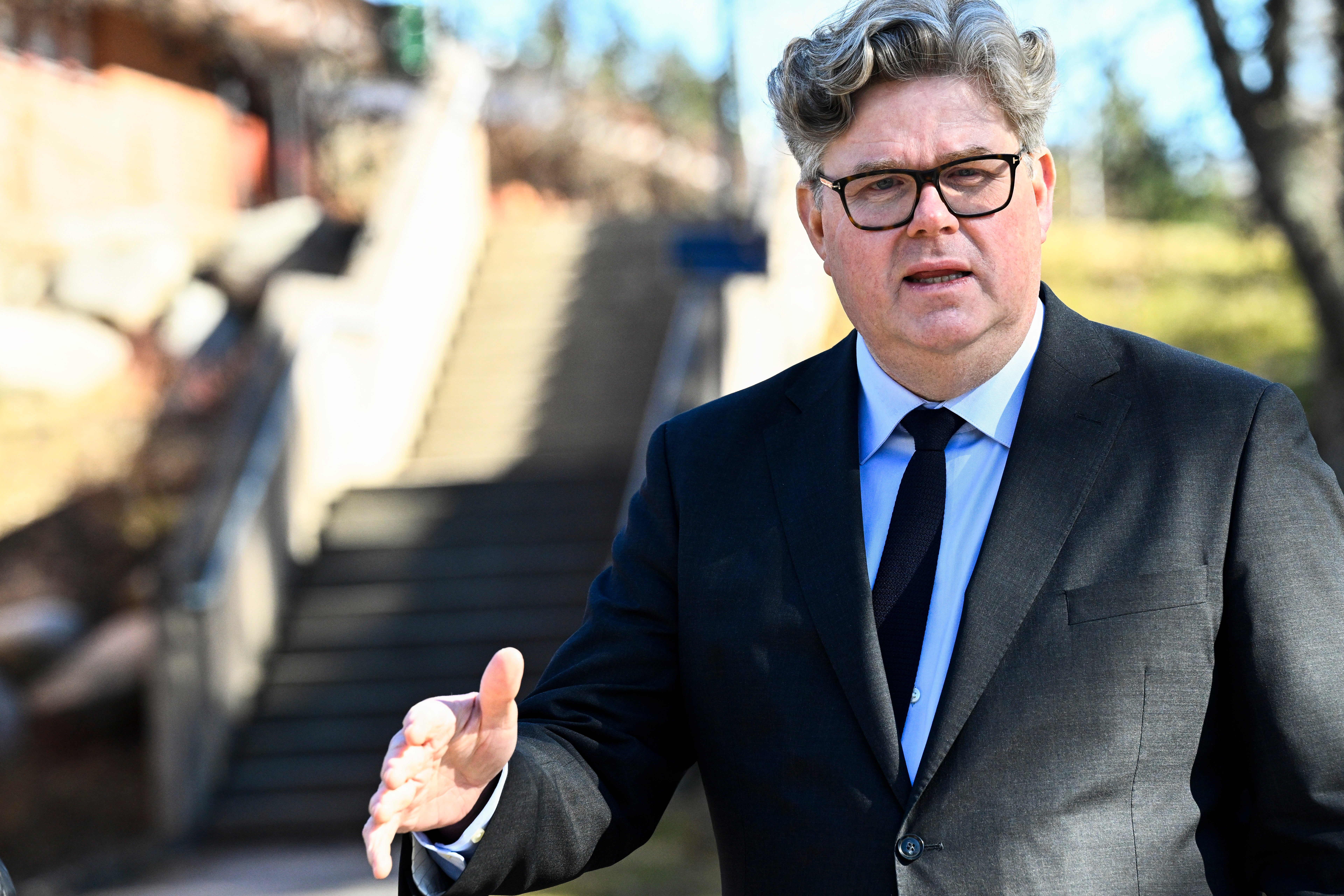 Man i kostym och glasögon står utomhus och gestikulerar medan han talar.