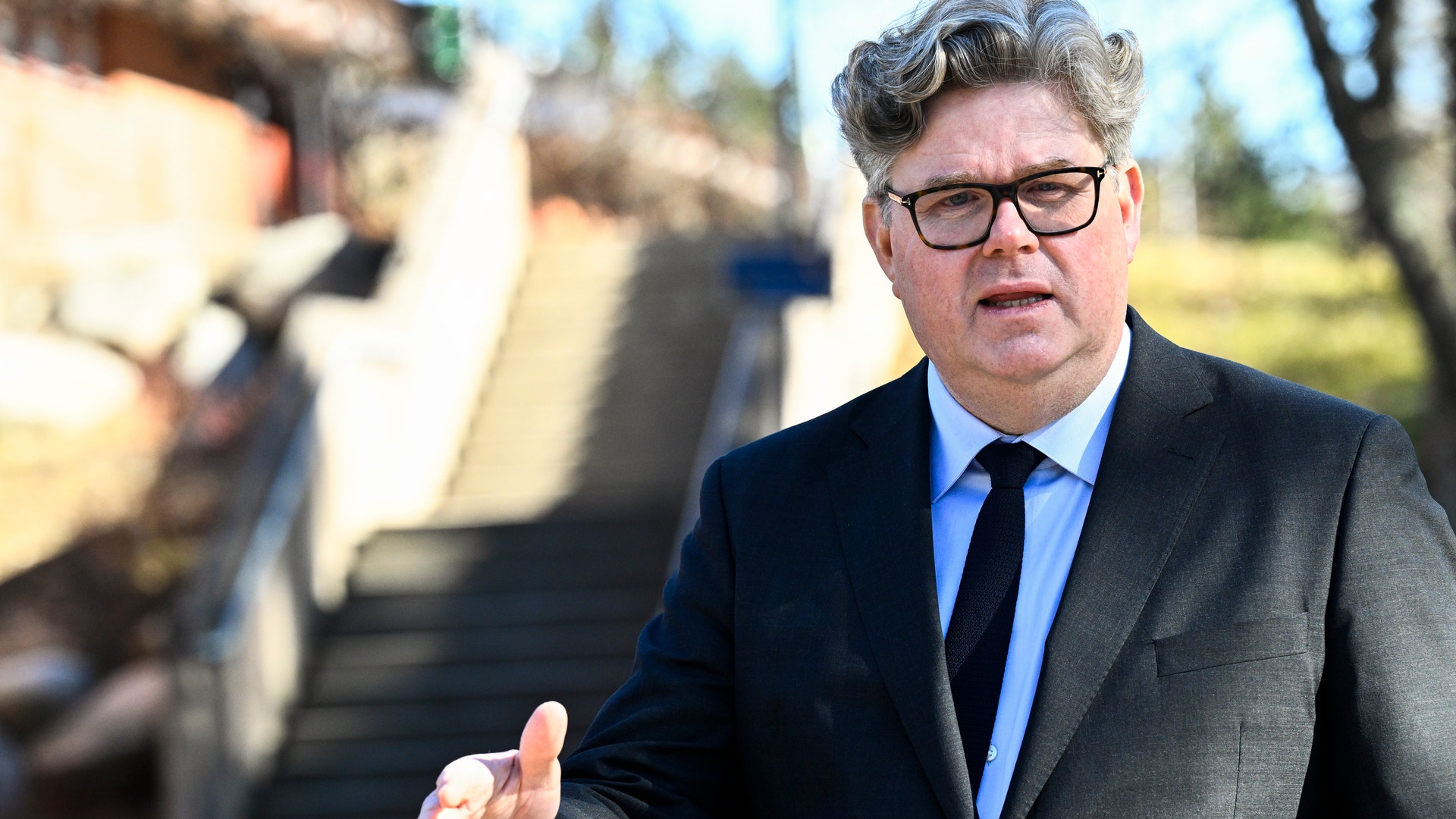 Man i kostym och glasögon står utomhus och gestikulerar medan han talar.