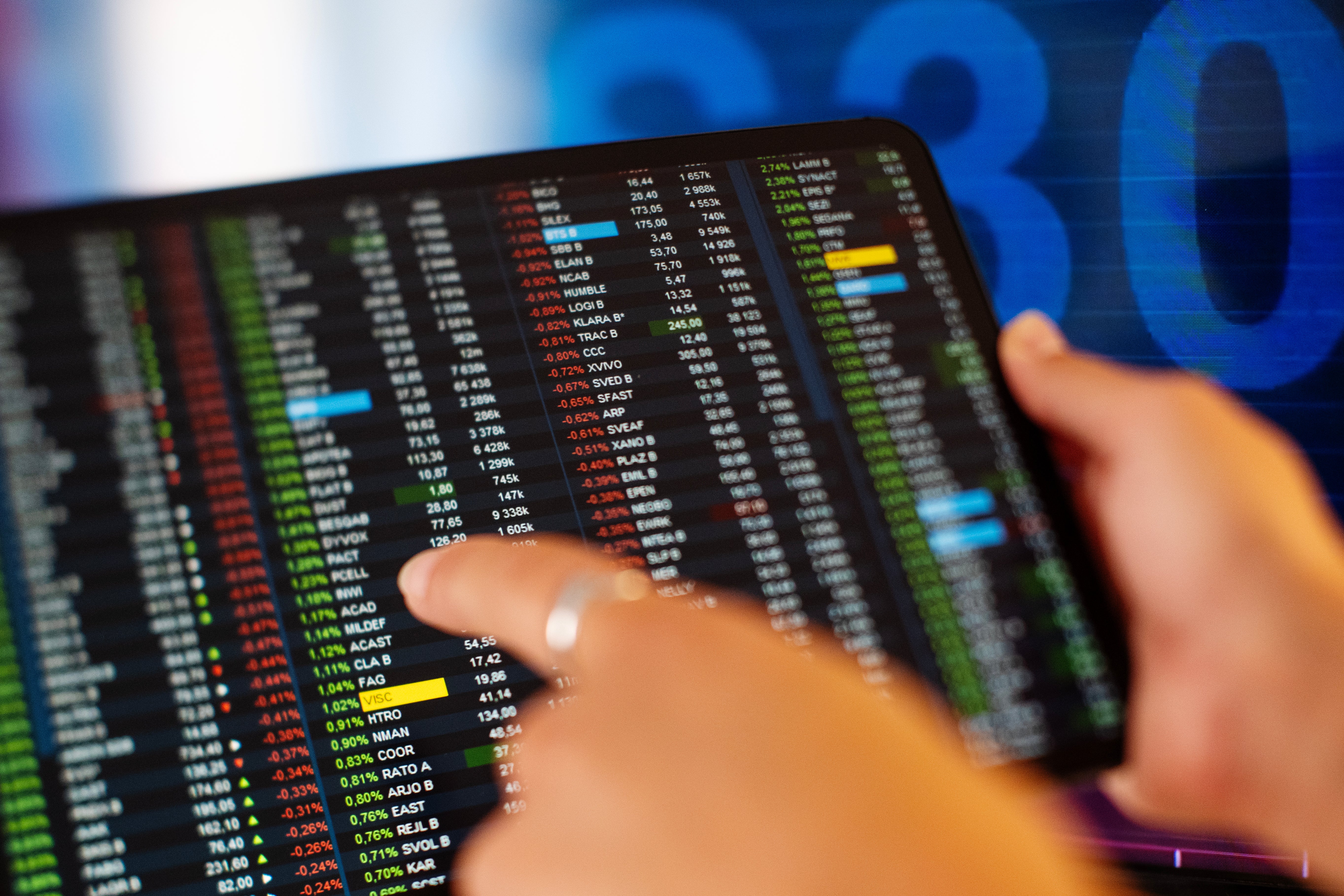 En person pekar på en skärm som visar finansiell data med aktiekurser och marknadsgrafer i olika färger.