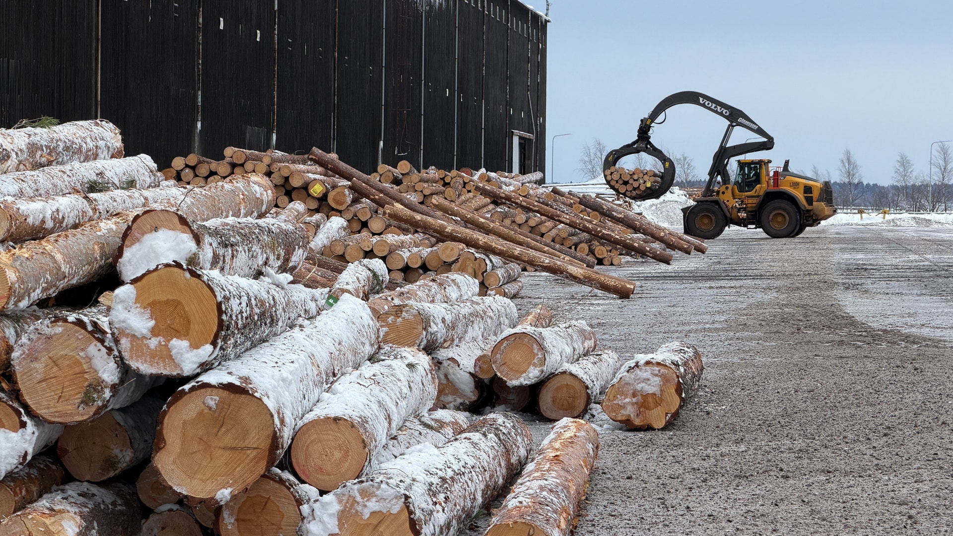 Timmerupplag på en snöig vintergård med staplad ved och skogsmaskiner