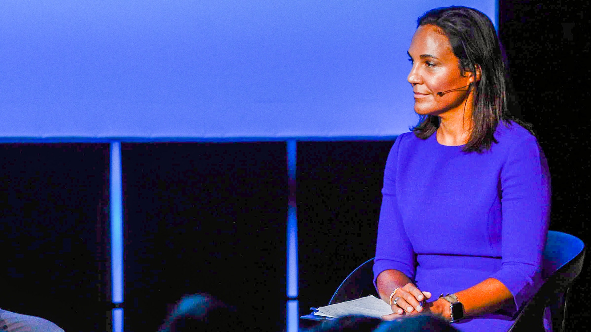 Kvinna i blå tröja talar på en scen med mikrofon headset.