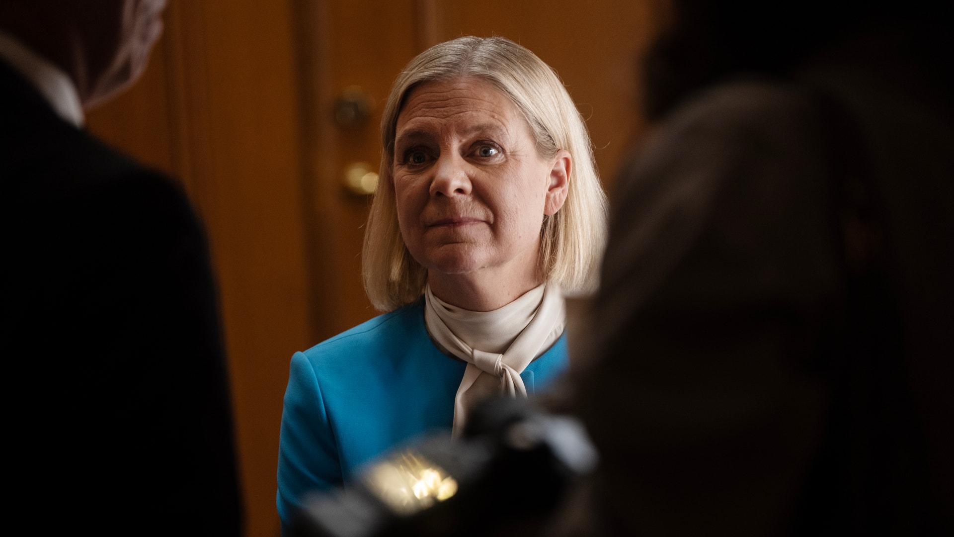 En kvinna i blå kavaj och sjal omgiven av personer i en korridor.