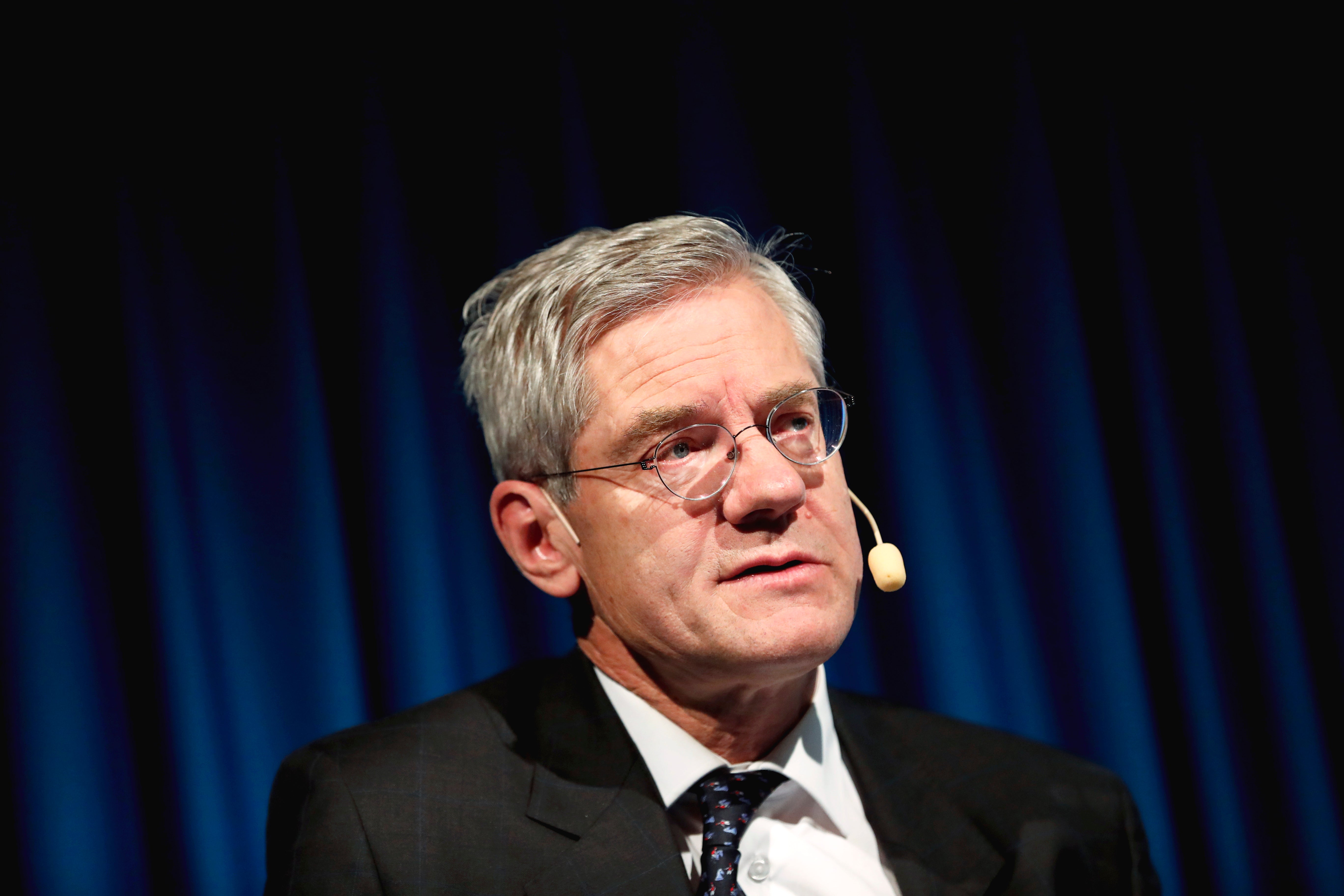 En äldre man i glasögon och formell svart kostym talar vid en scen framför blå gardiner.