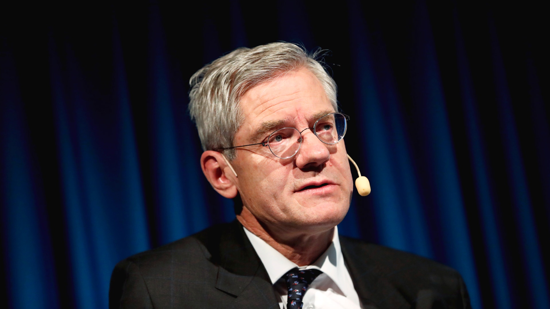 En äldre man i glasögon och formell svart kostym talar vid en scen framför blå gardiner.