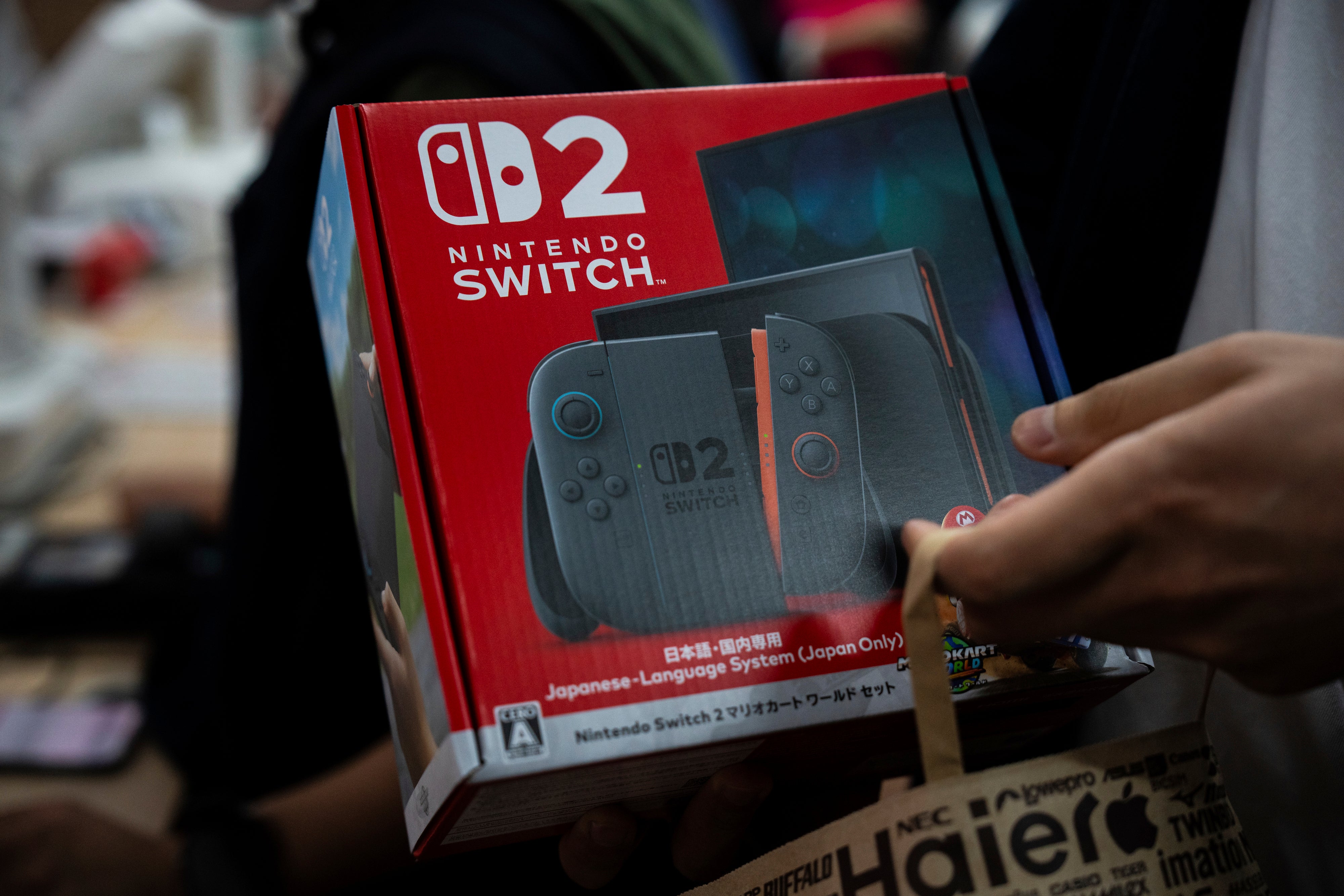 En person håller upp en röd Nintendo Switch 2-förpackning med spelkontroller synliga på boxen.
