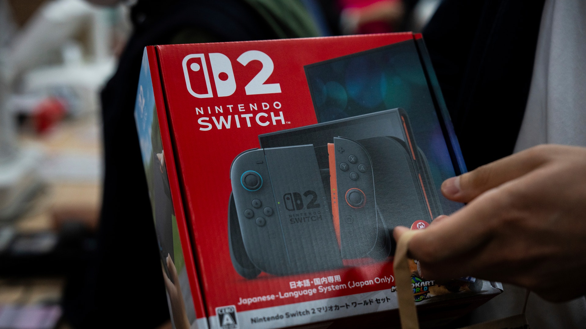 En person håller upp en röd Nintendo Switch 2-förpackning med spelkontroller synliga på boxen.