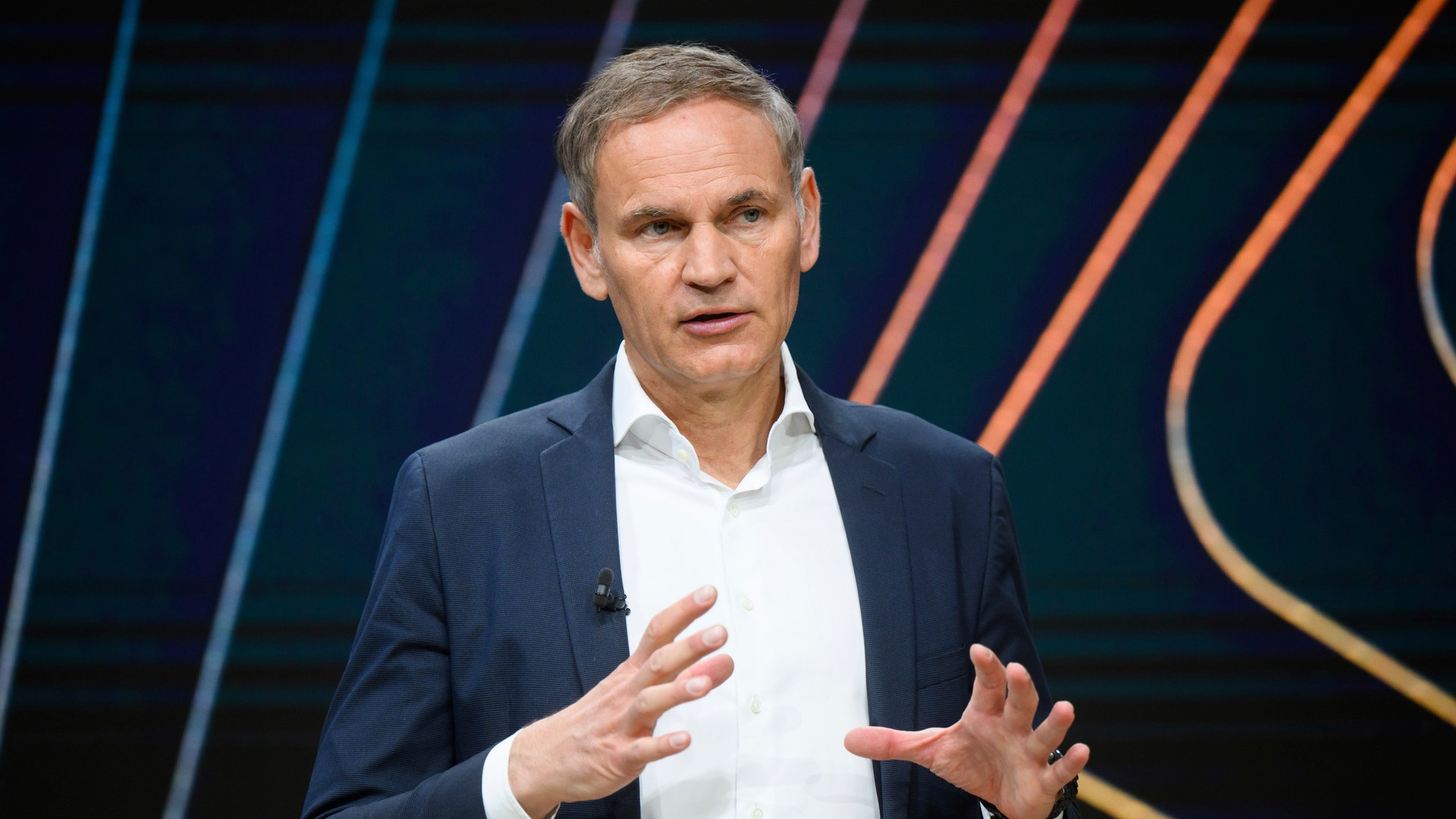 Man i mörk kostym och vit skjorta håller presentation på scen med färgad bakgrund