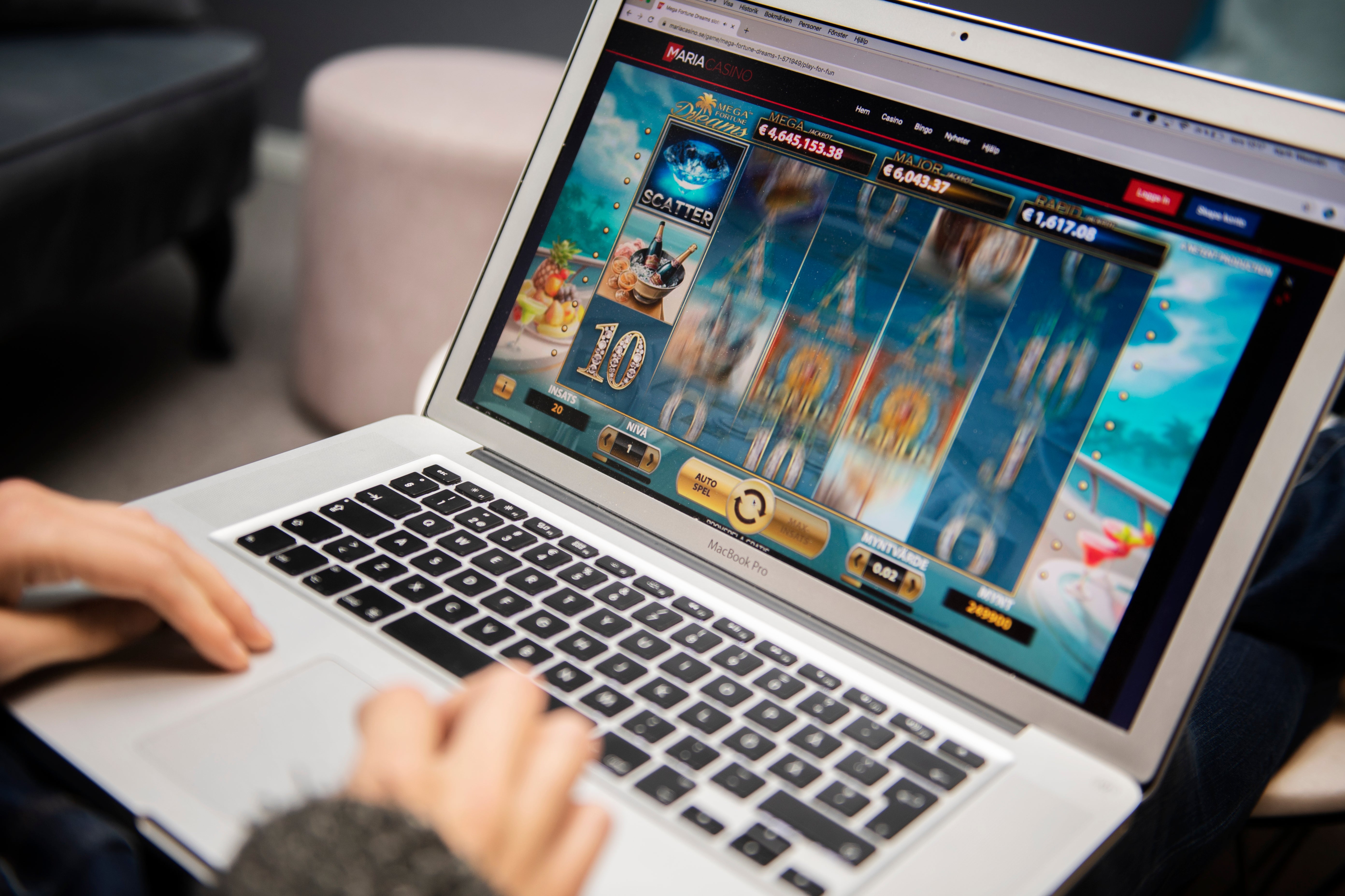 Person spelar ett onlinespel på en laptop. Skärmen visar ett slotspel med olika symboler och en blå himmelsbakgrund.