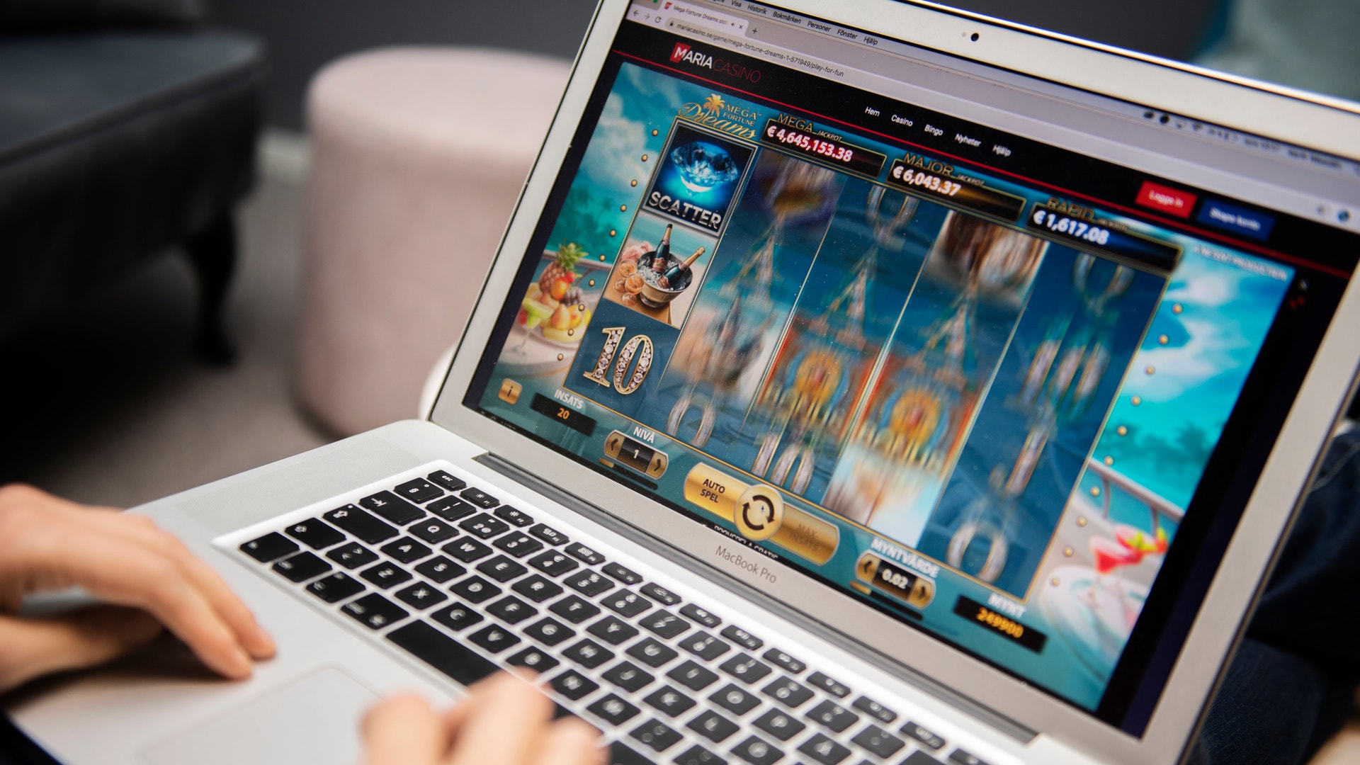 Person spelar ett onlinespel på en laptop. Skärmen visar ett slotspel med olika symboler och en blå himmelsbakgrund.