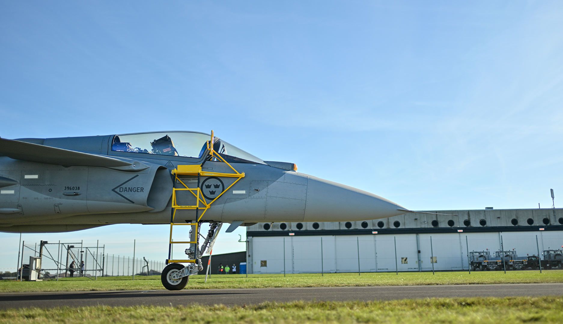 Gripen E stridsflygplan parkerat på flygfält med gul servicestege framför cockpiten, hangar i bakgrunden