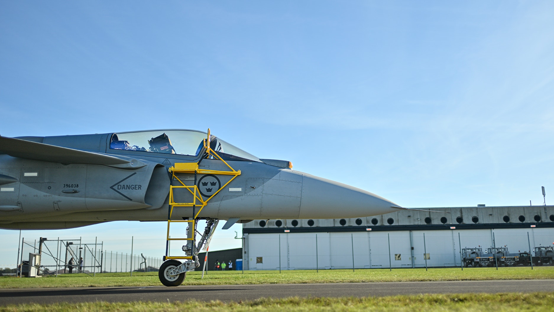 Gripen E stridsflygplan parkerat på flygfält med gul servicestege framför cockpiten, hangar i bakgrunden