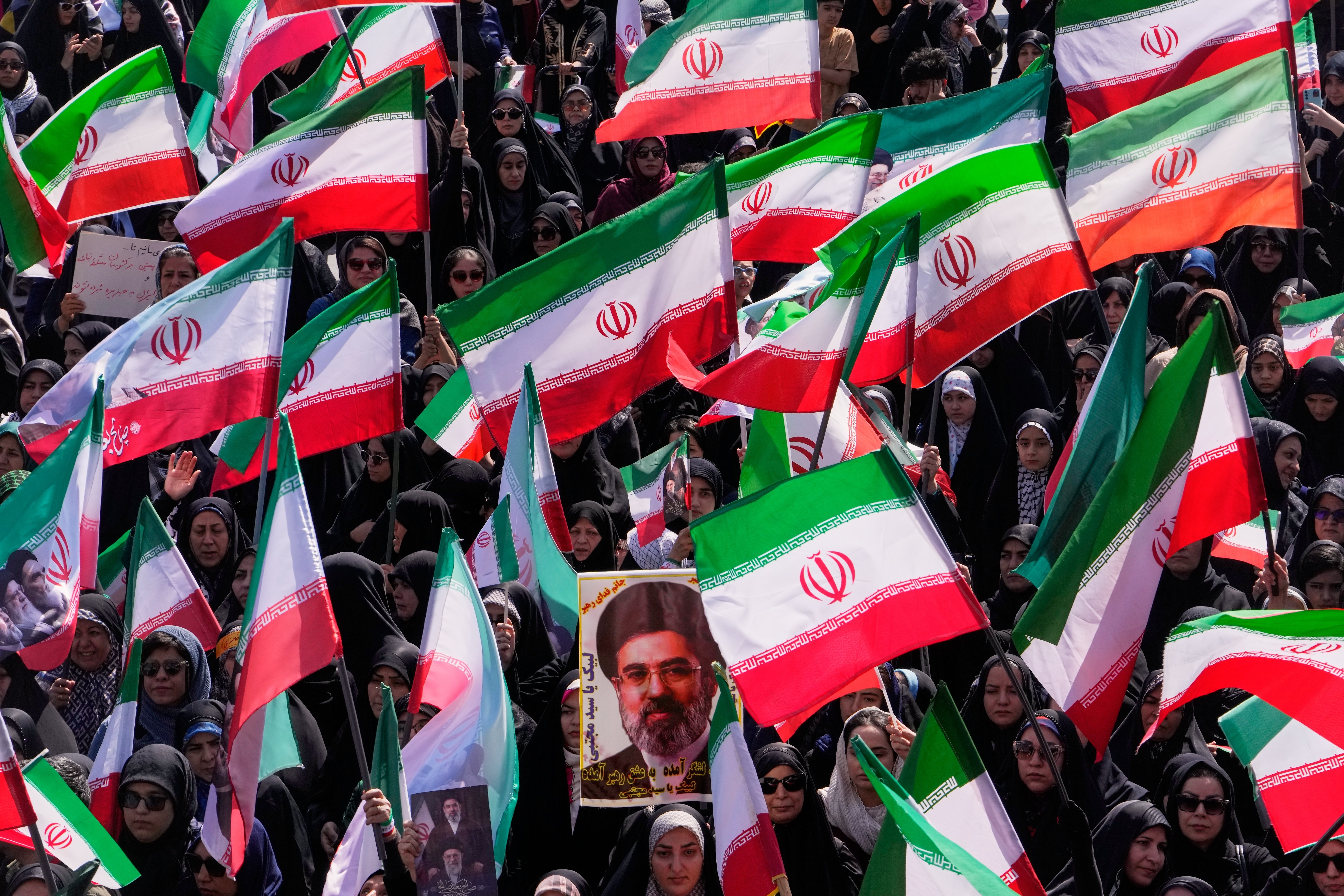 Stor folksamling med människor som håller upp iranska flaggor. En porträttbild av en man är synlig bland flaggorna.