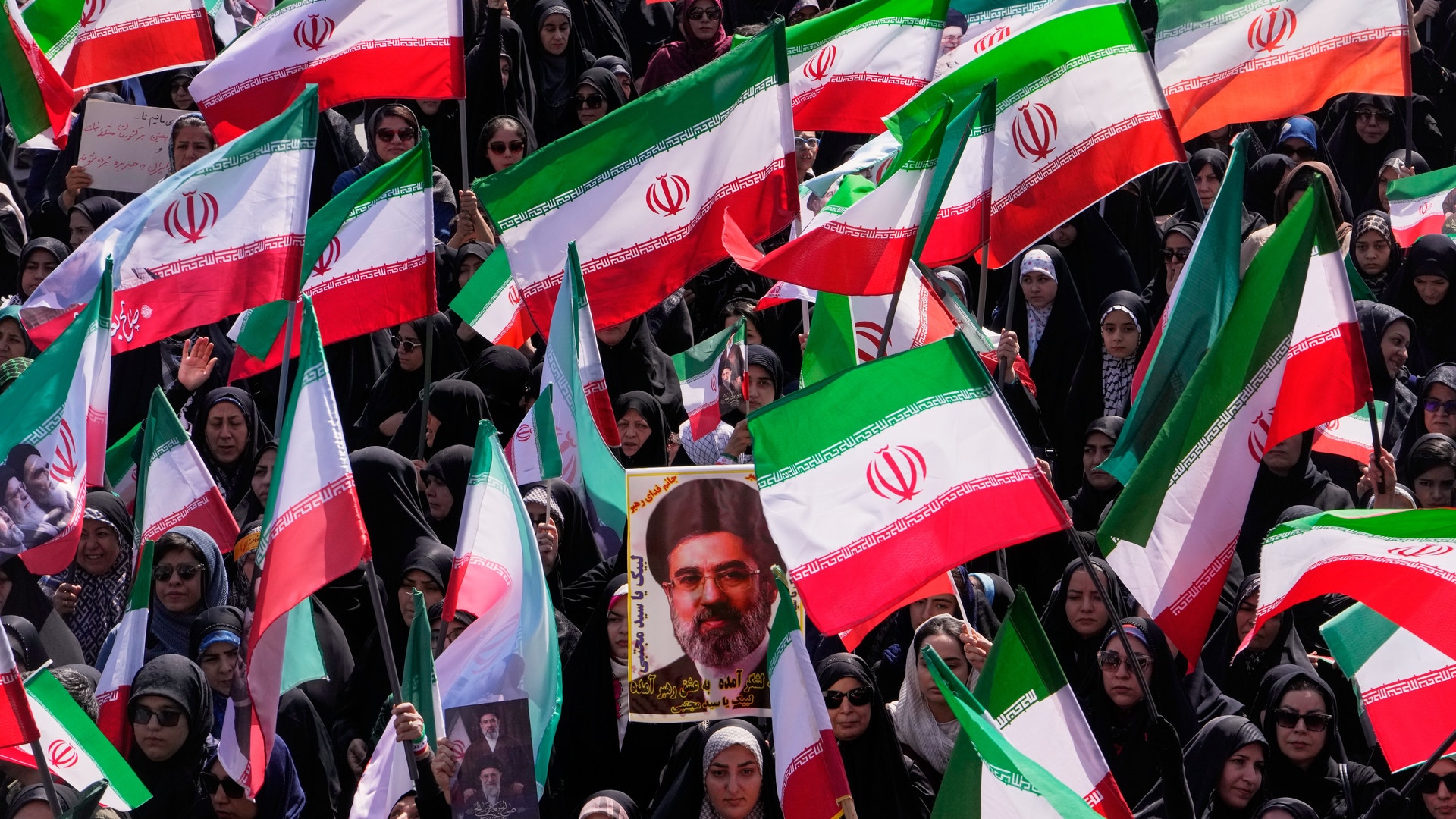 Stor folksamling med människor som håller upp iranska flaggor. En porträttbild av en man är synlig bland flaggorna.