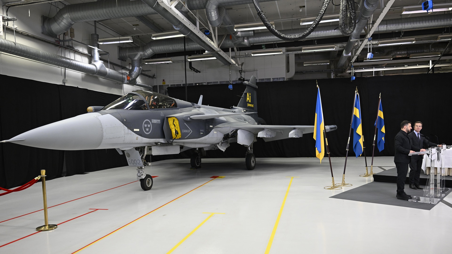 Ett svenskt stridsflygplan Gripen står i ett hangar med svenska flaggor i bakgrunden. Två personer står bredvid flygplanet.