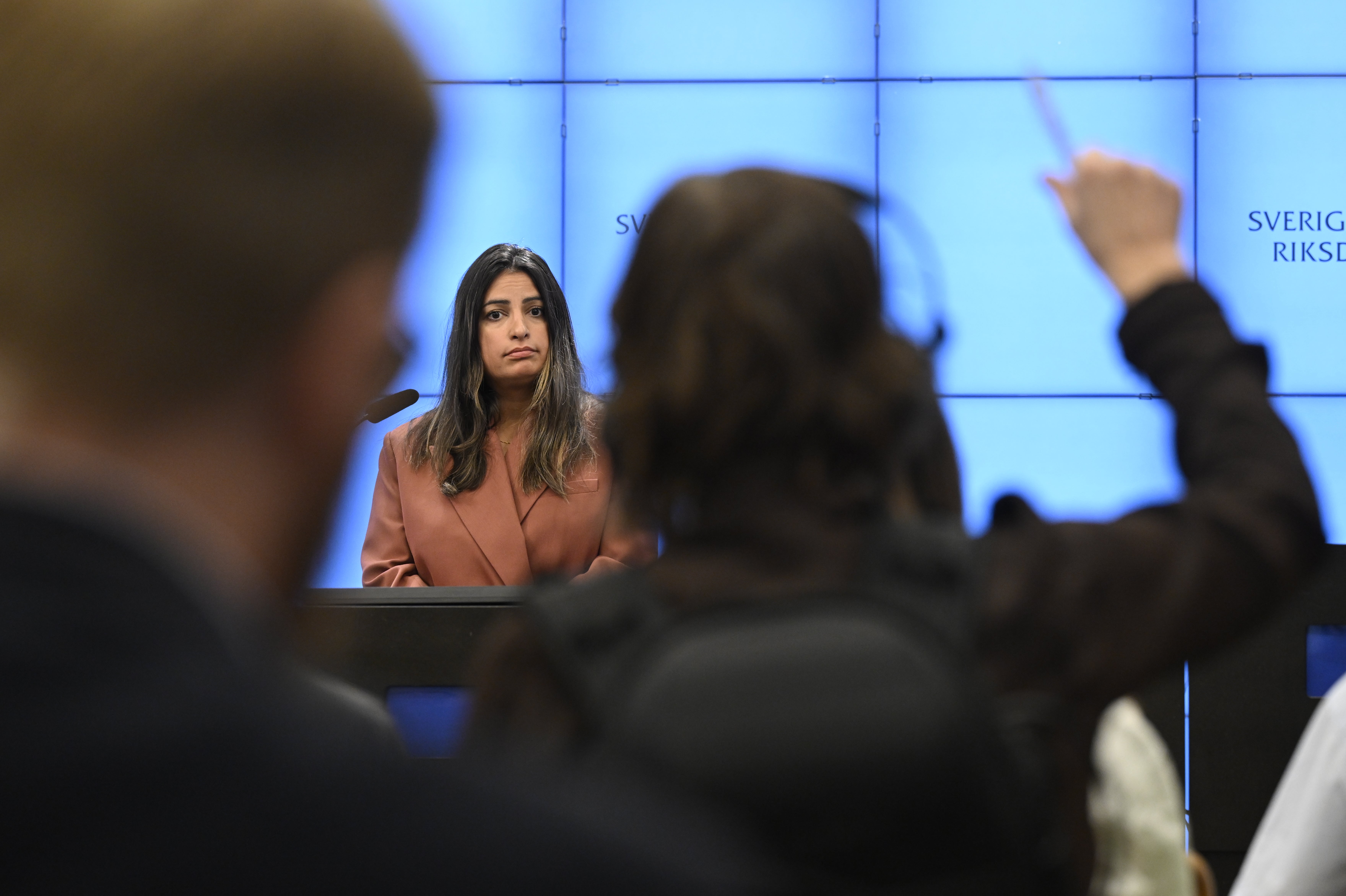 Pressträff i Sveriges riksdag där en kvinna talar vid ett podium framför journalister, en journalist räcker upp handen för att ställa en fråga.