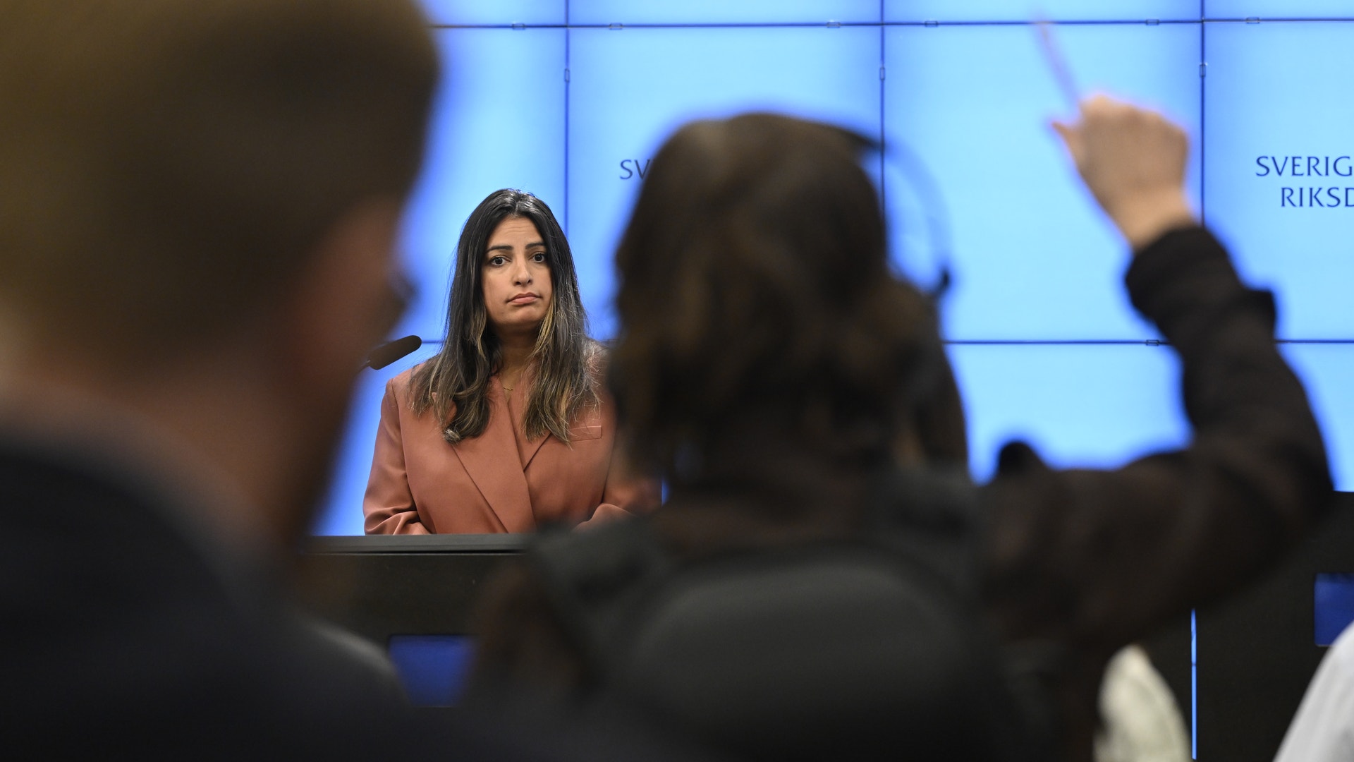 Pressträff i Sveriges riksdag där en kvinna talar vid ett podium framför journalister, en journalist räcker upp handen för att ställa en fråga.