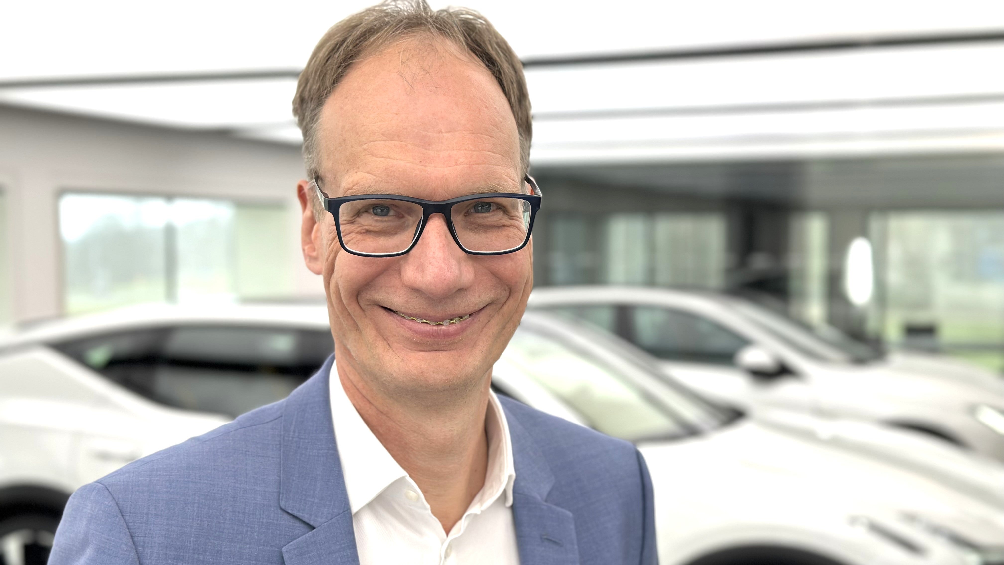Porträttfoto av en man med glasögon och blå kavaj som ler mot kameran. I bakgrunden syns en modern garage med vita bilar.