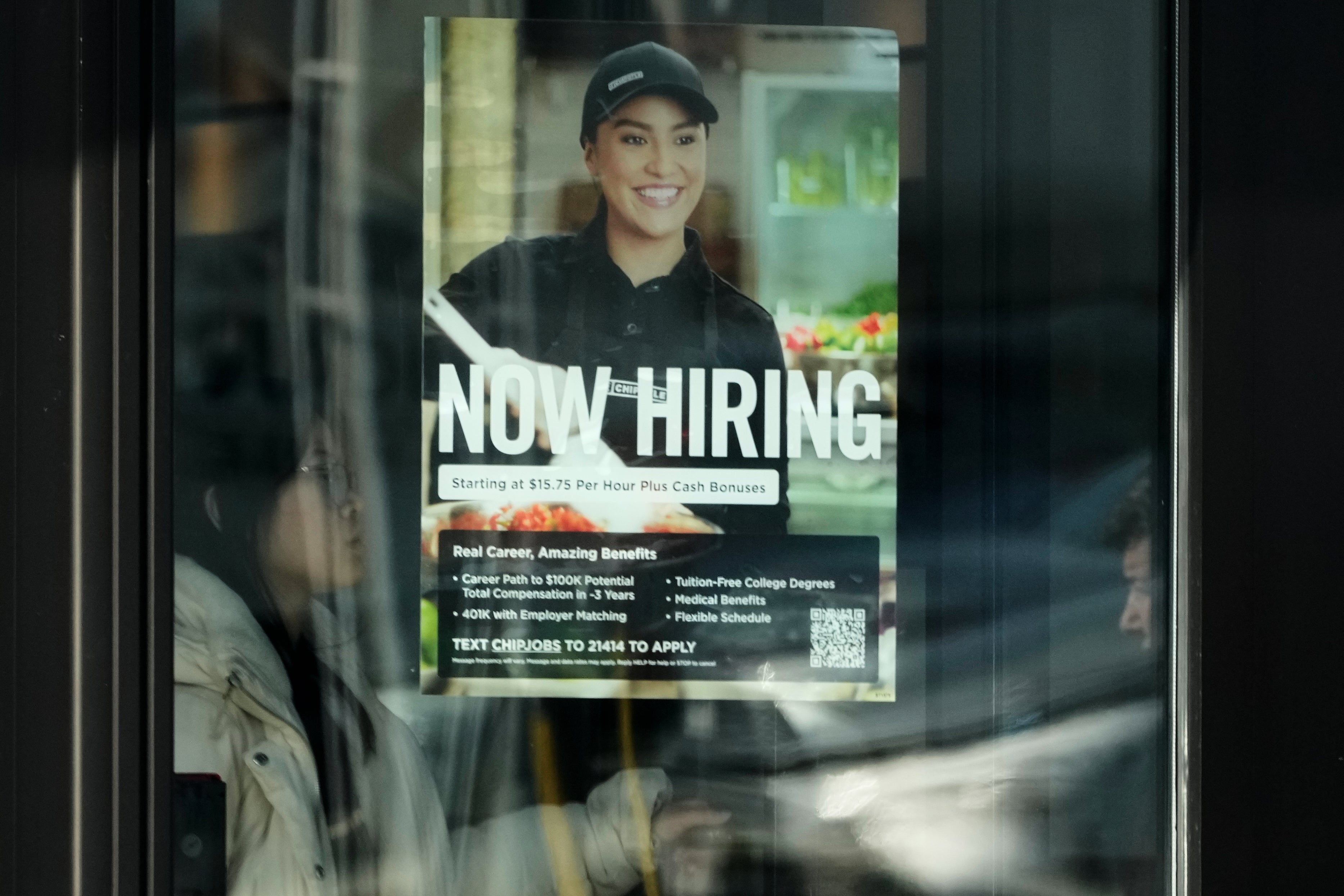Anställningsaffisch på glasfönster med texten "NOW HIRING". Affischen visar en leende person i svart uniform och keps, med information om lön, förmåner och anvisningar för att söka jobb via SMS.