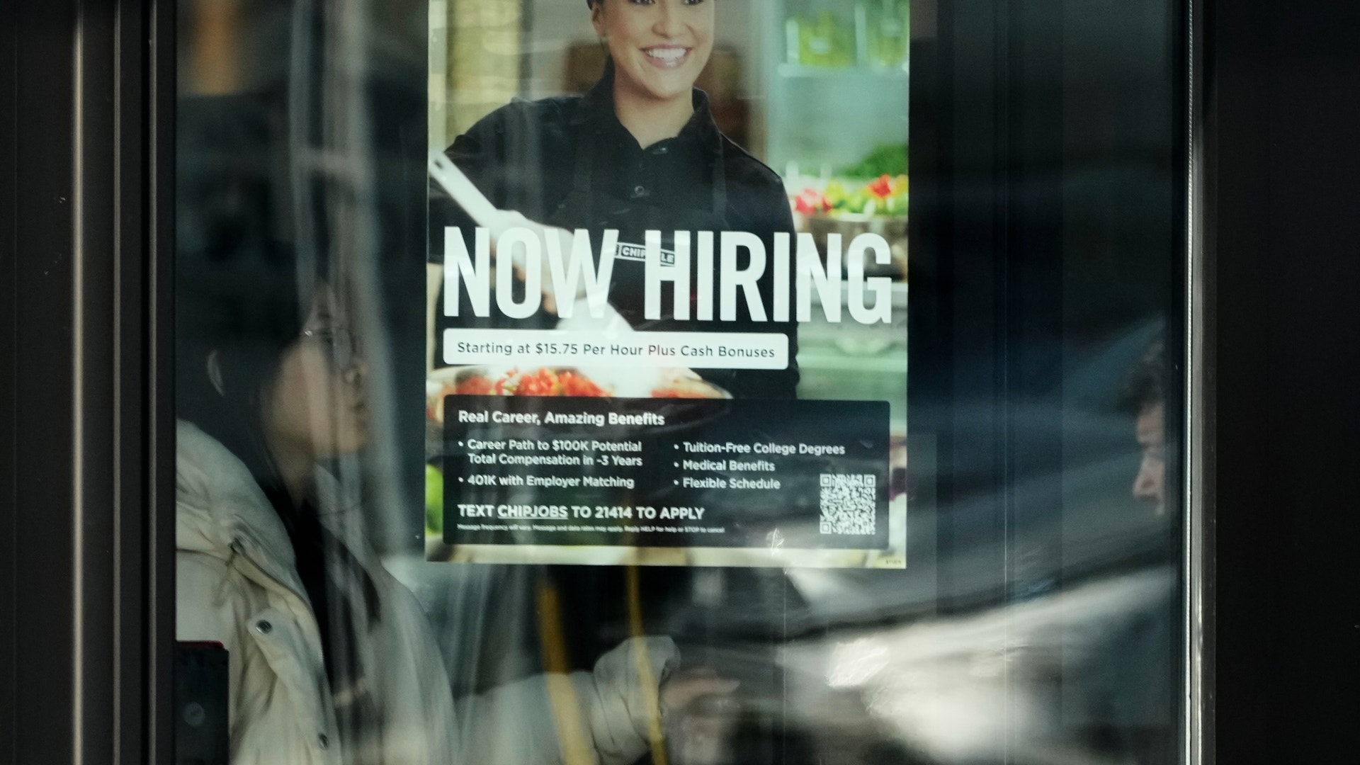Anställningsaffisch på glasfönster med texten "NOW HIRING". Affischen visar en leende person i svart uniform och keps, med information om lön, förmåner och anvisningar för att söka jobb via SMS.