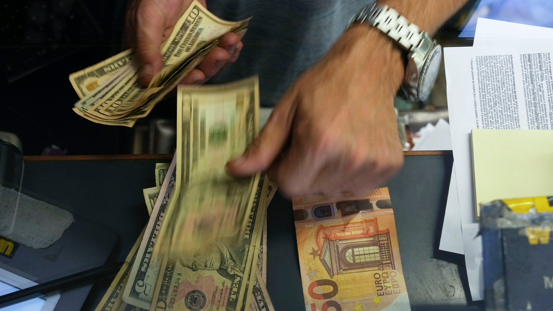 Två händer hanterar amerikanska dollar och eurosedlar på ett skrivbord med papper.