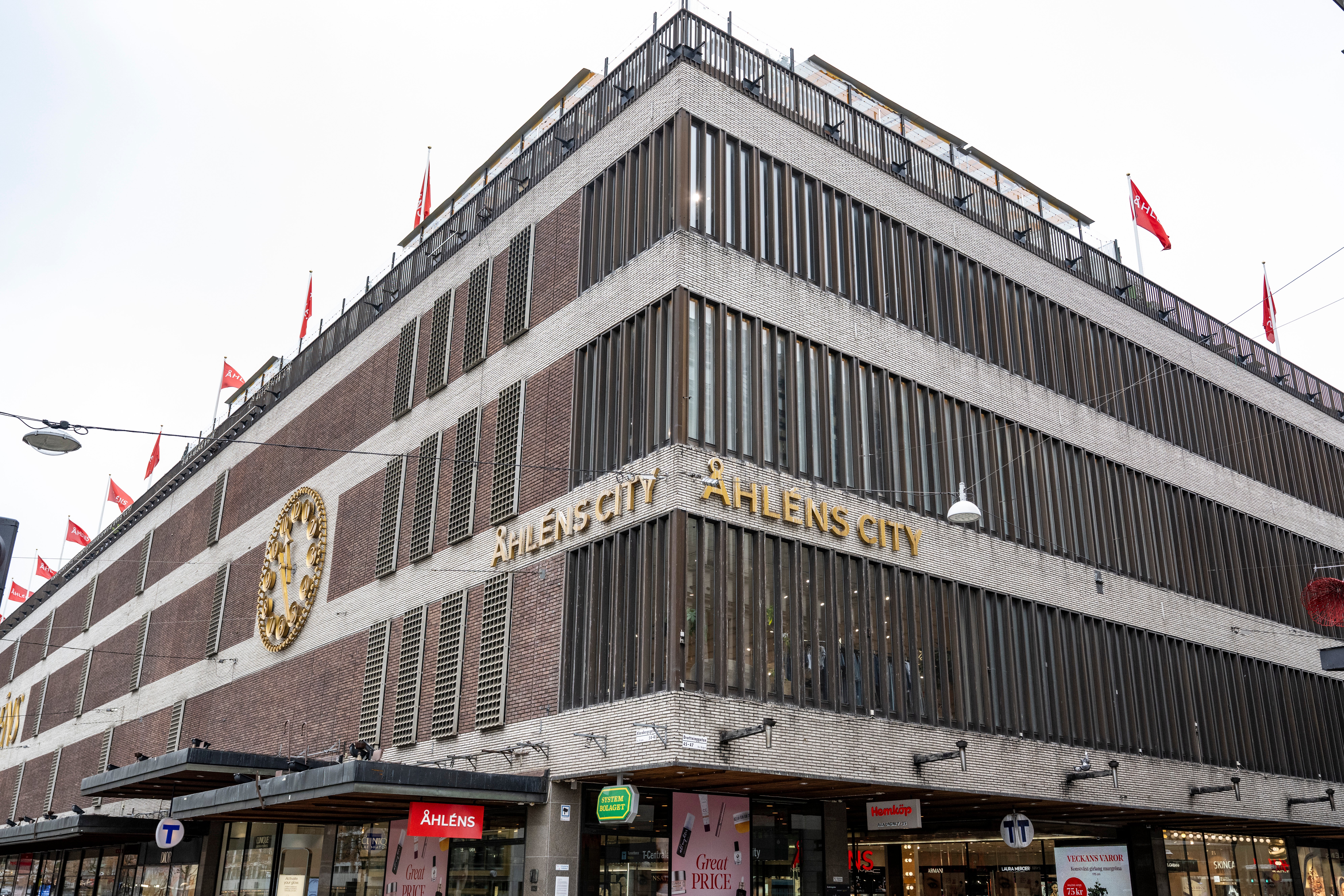 En stor modernistisk byggnad med tegelstenar och betong, flera våningar med horisontella linjer och röda flaggor på taket. Skyltfönster och butiker på bottenvåningen.