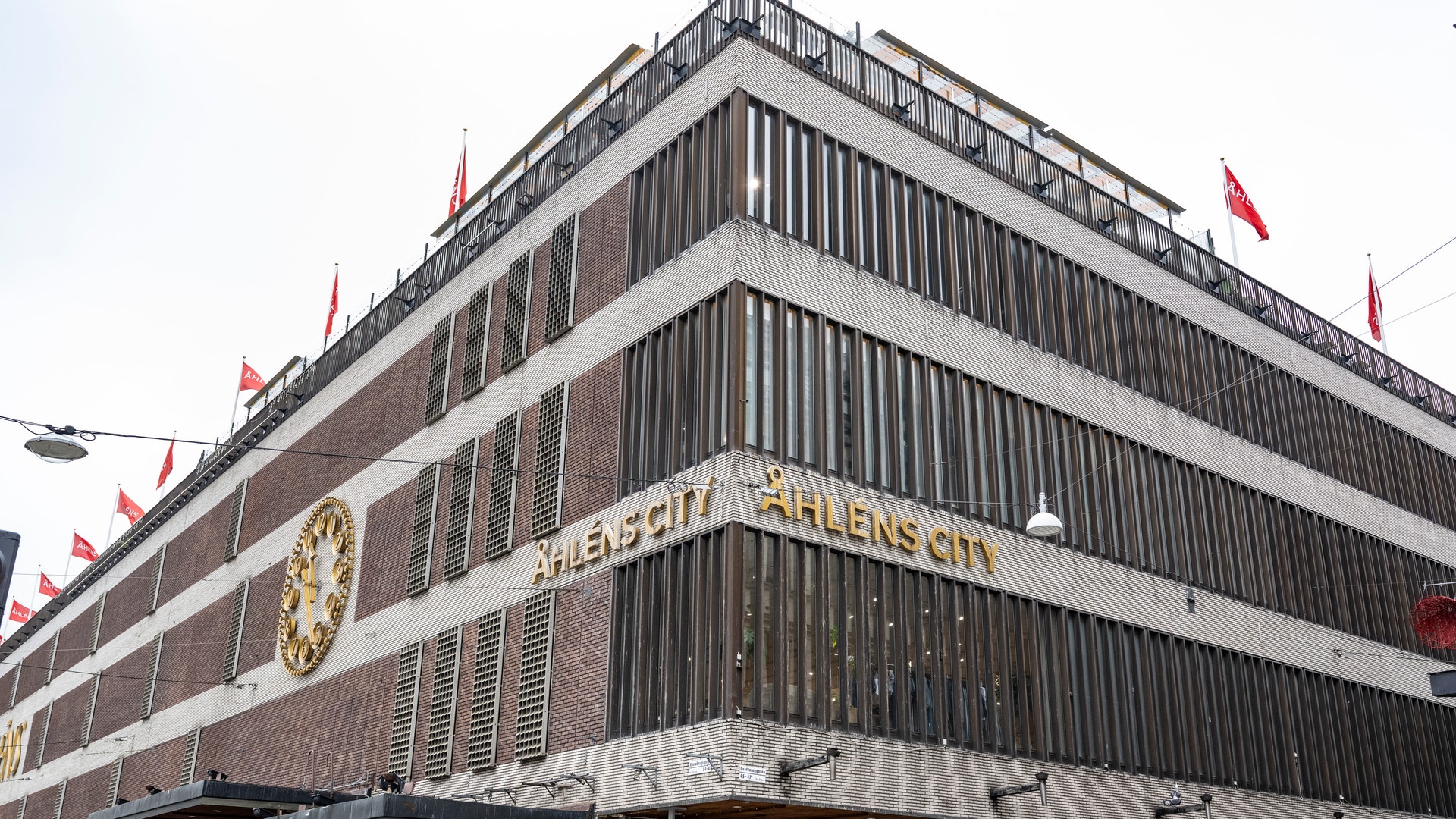 En stor modernistisk byggnad med tegelstenar och betong, flera våningar med horisontella linjer och röda flaggor på taket. Skyltfönster och butiker på bottenvåningen.