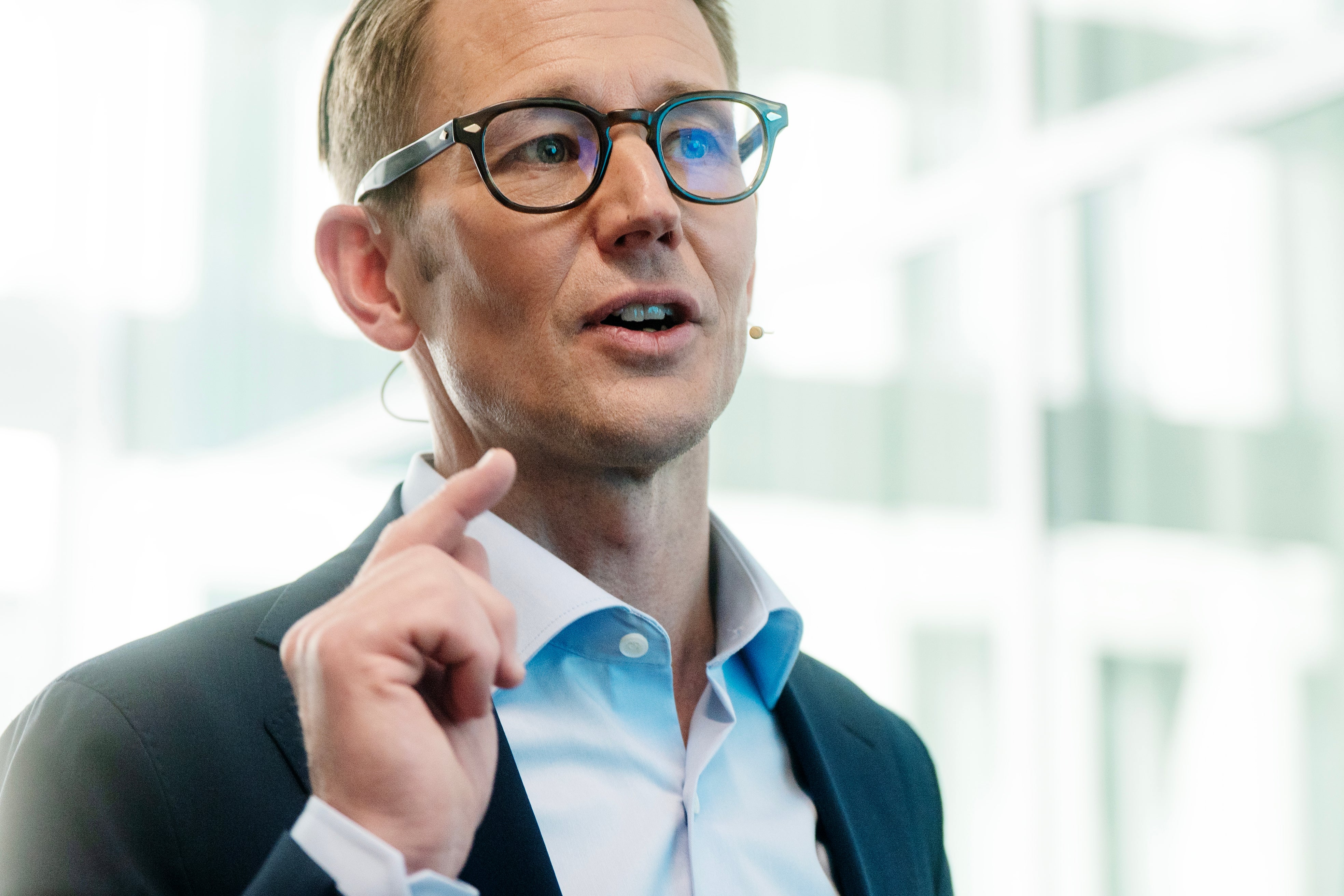 Man med glasögon i blå skjorta och kavaj som håller ett tal framför ett modernt kontorslandskap.